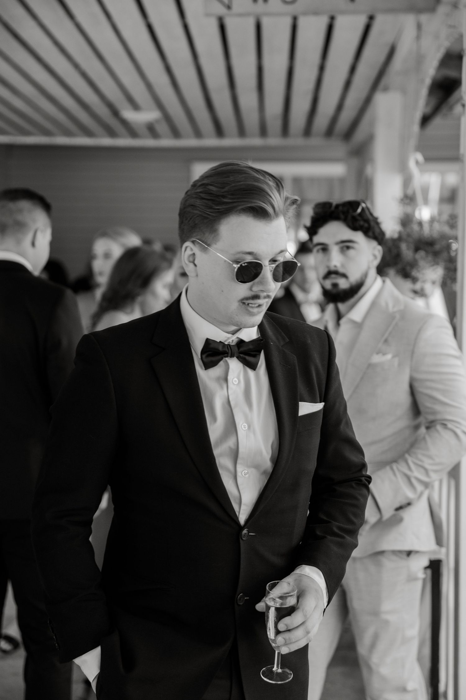 Stylish wedding guest in a black tuxedo and sunglasses holding a champagne glass at an outdoor reception.