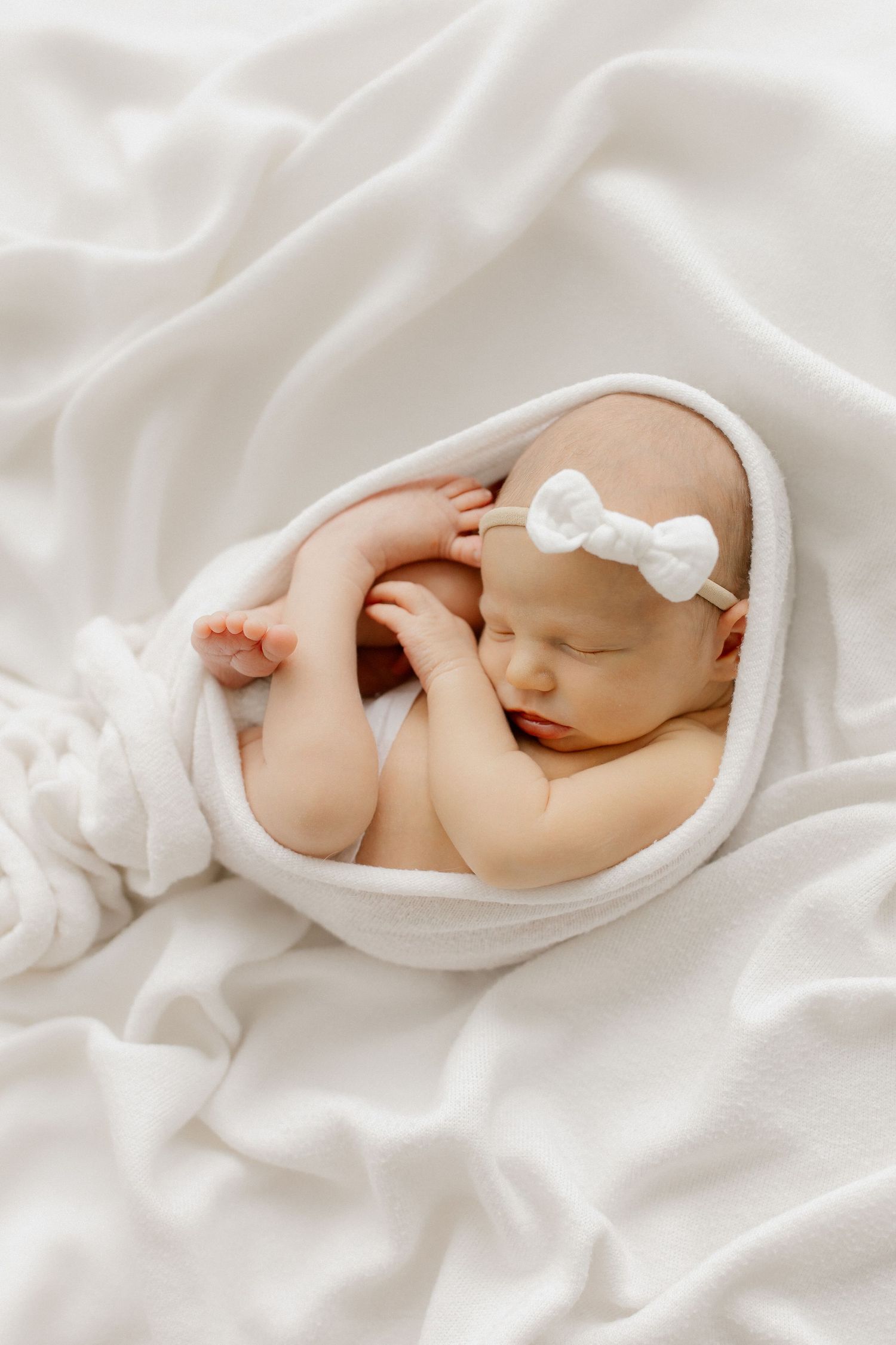 Newborn baby girl sleeping peacefully wrapped in a white blanket with a white bow headband.
