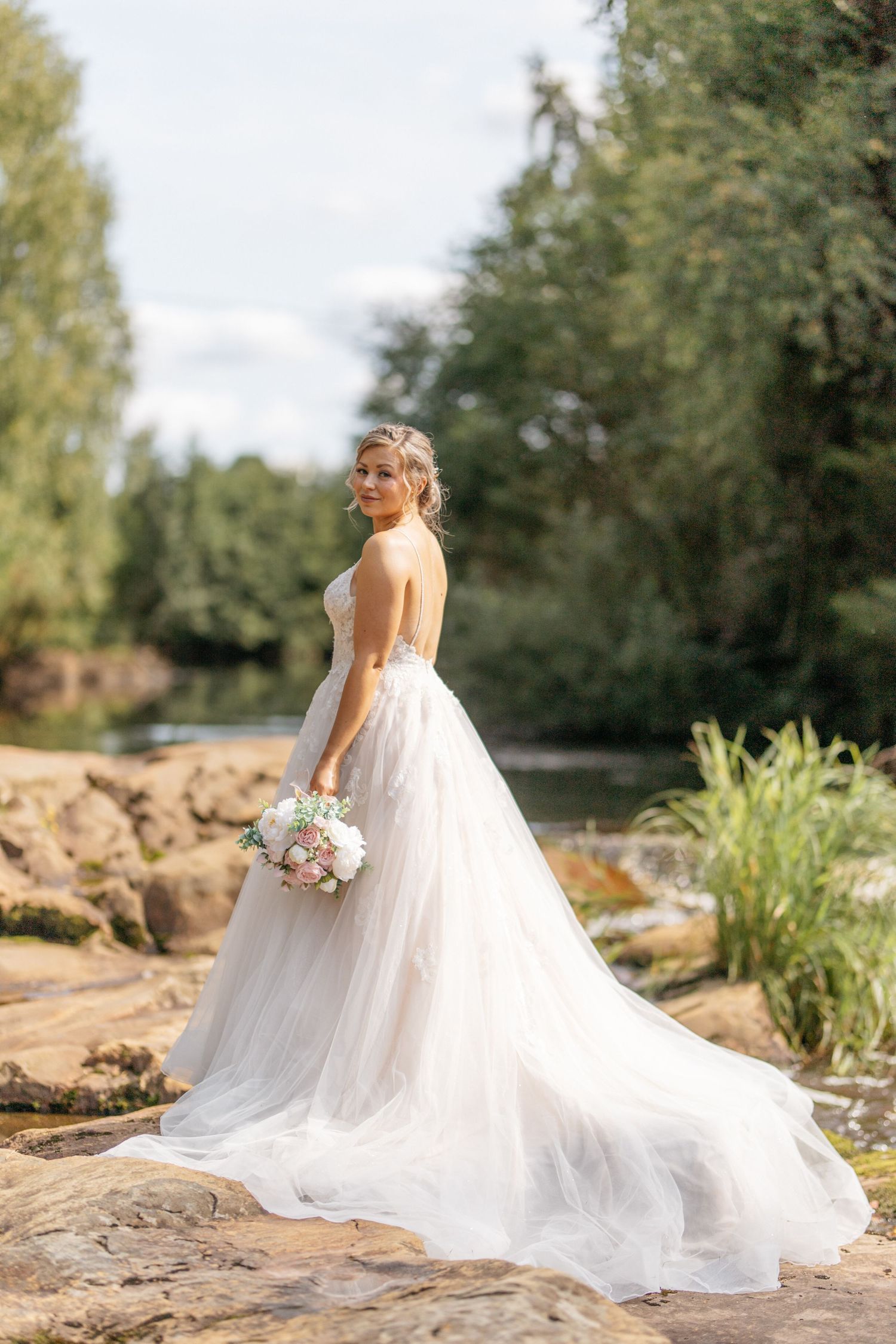 Bride in flowing white gown poses with bouquet by scenic pond and rocks.