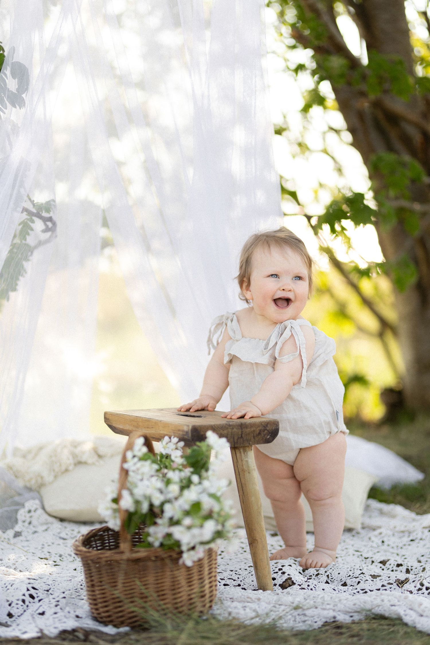 Baby in white romper laughs while standing at wooden stool with floral basket nearby.