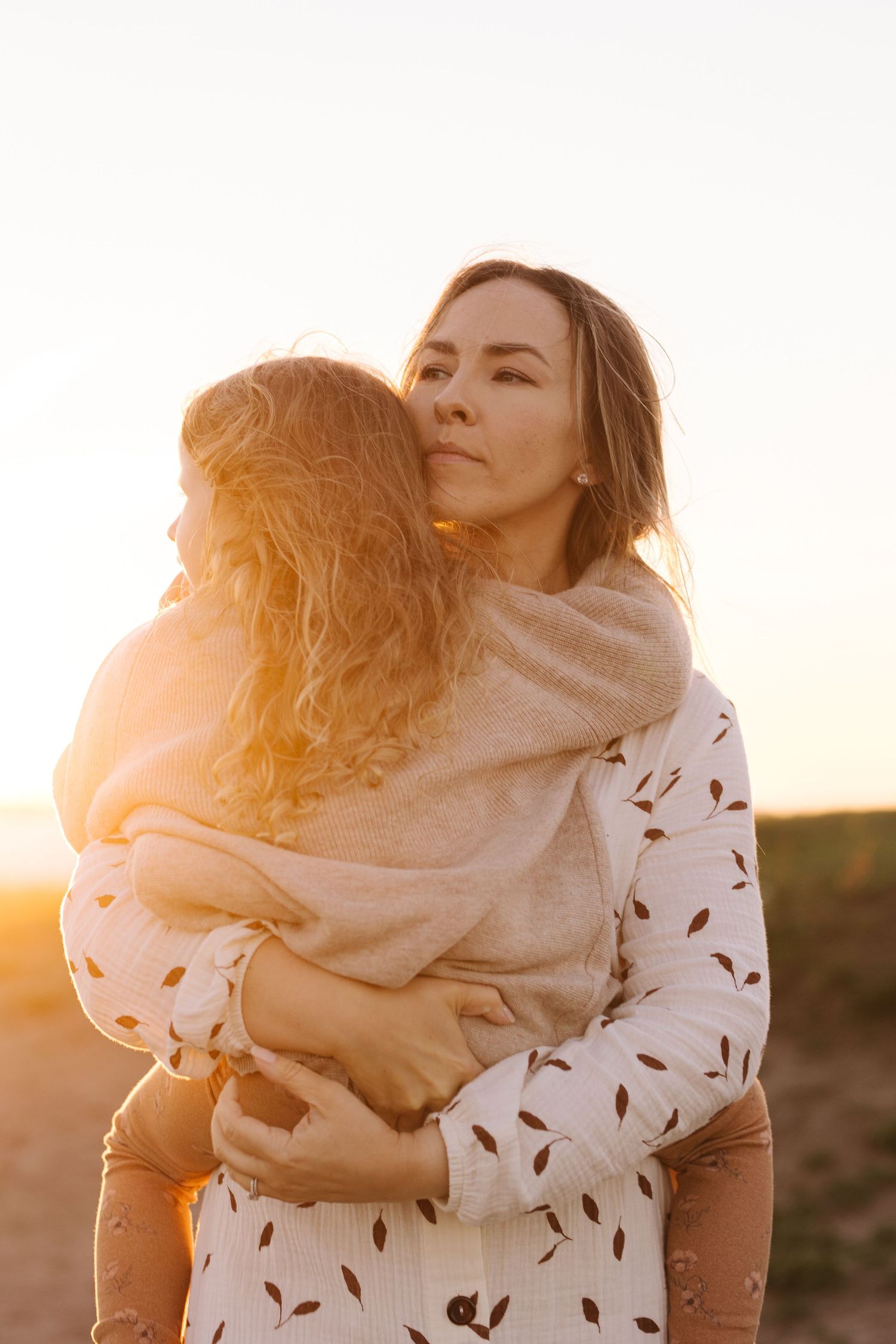 Mother and child embrace at sunset in white dress with flowing shawl during golden hour photoshoot.
