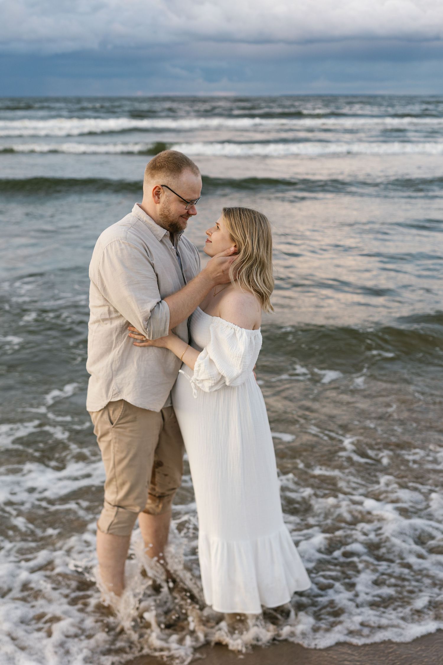 A romantic couple embraces on a beach at sunset as ocean waves lap at their feet.