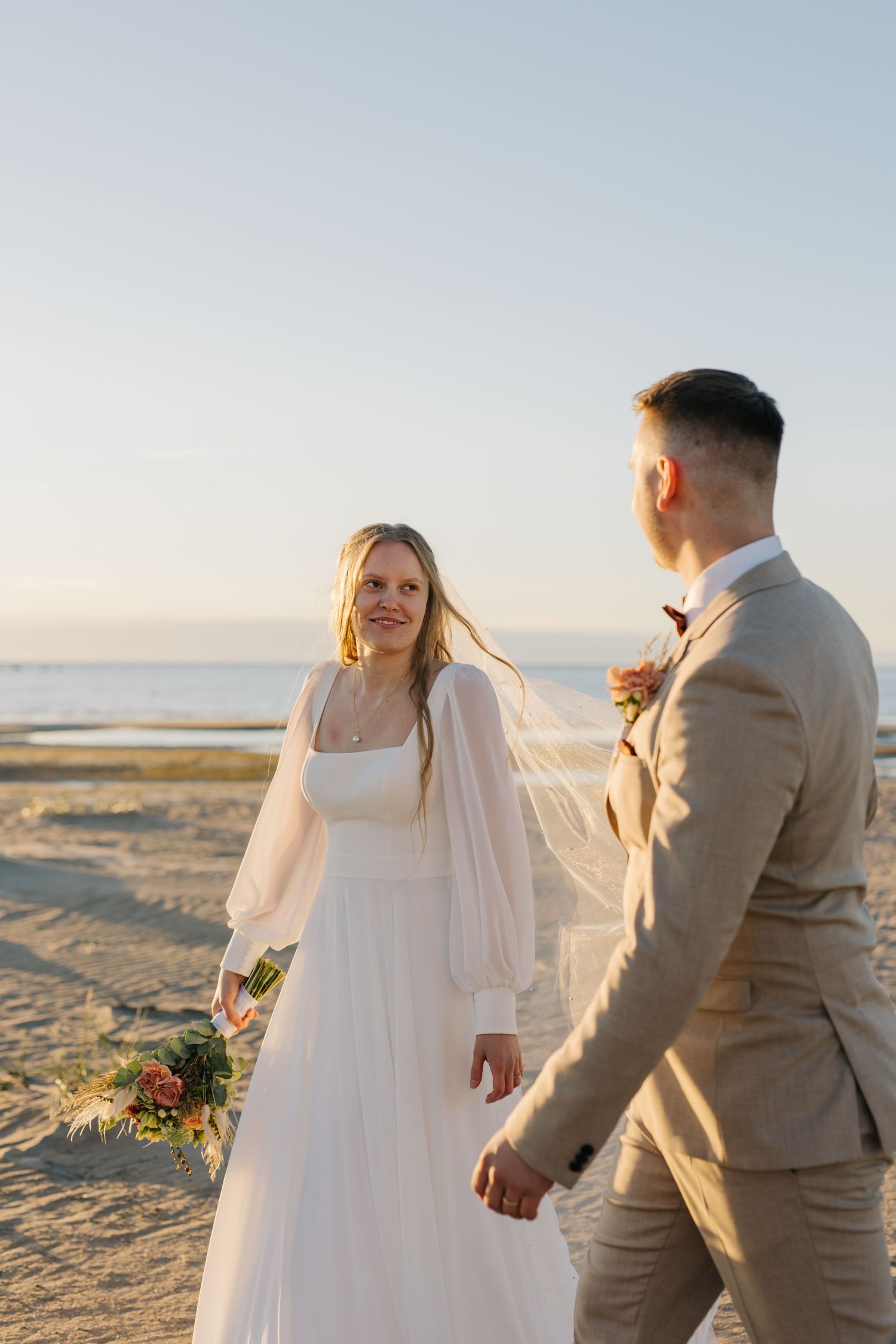 A couple in beachwear walks along a sandy shore at sunset, with the bride wearing a flowing white dress and cape.