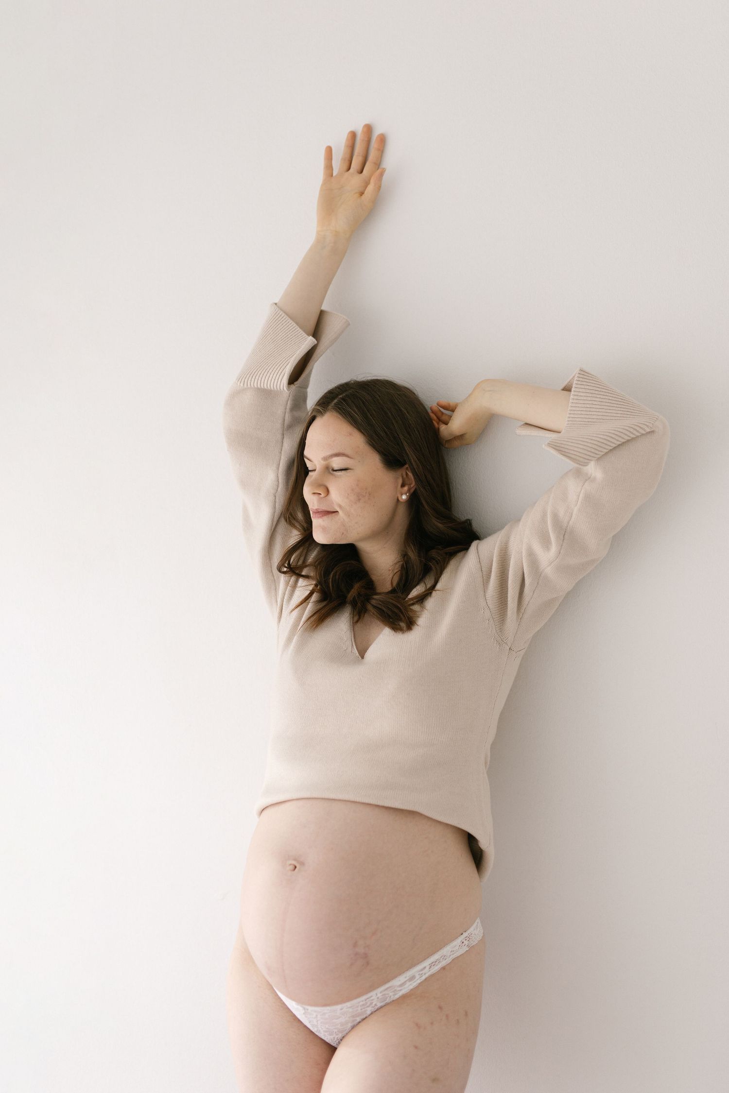 Pregnant woman in beige sweater poses against white wall with arms raised.