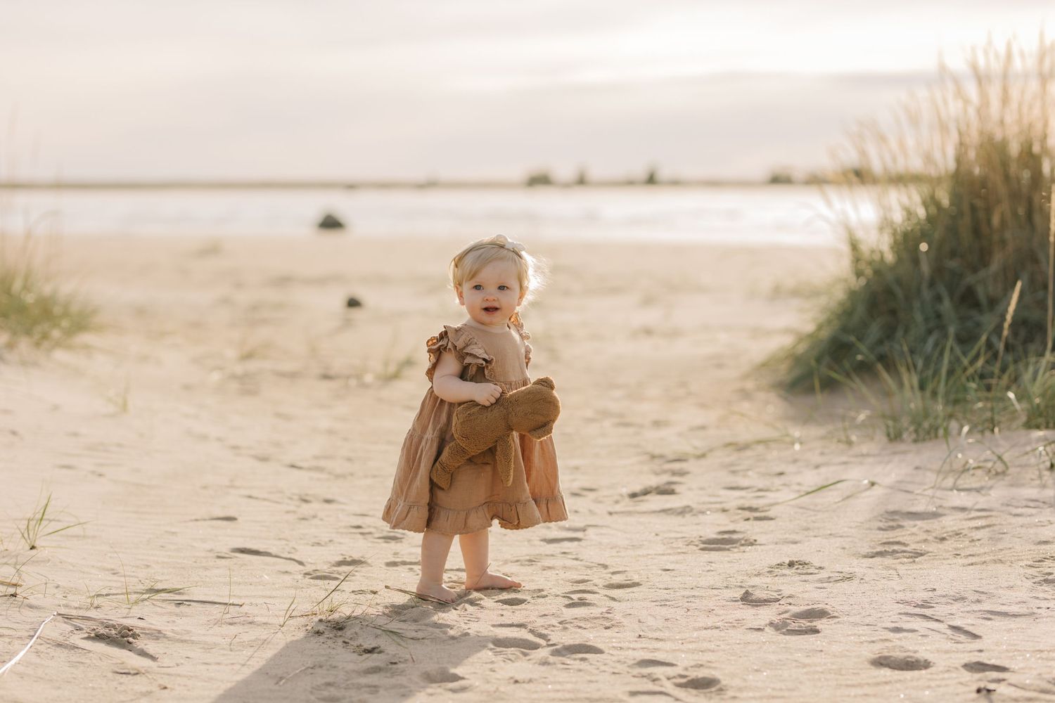 Small child wearing brown dress stands on sandy beach near grassy dunes at sunset.