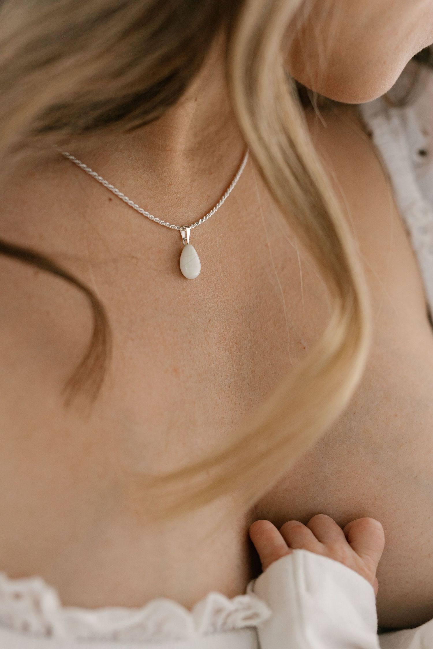 Close-up of delicate silver teardrop necklace against skin with baby's hand visible.