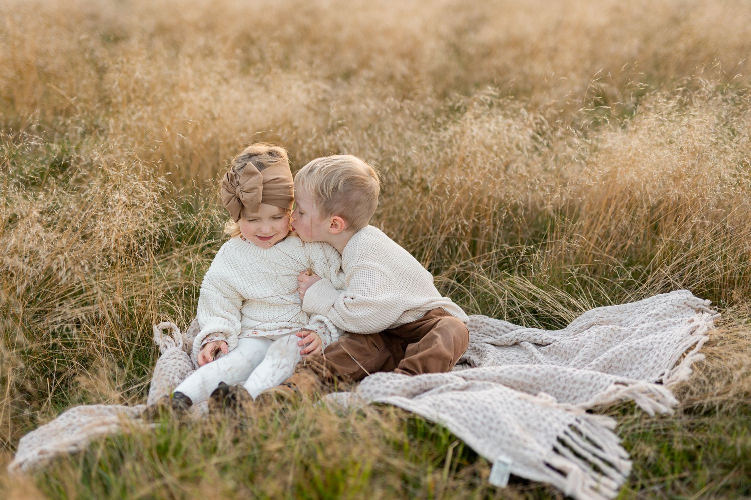 Two small children sit together on a white blanket in a sunlit meadow of tall grass.