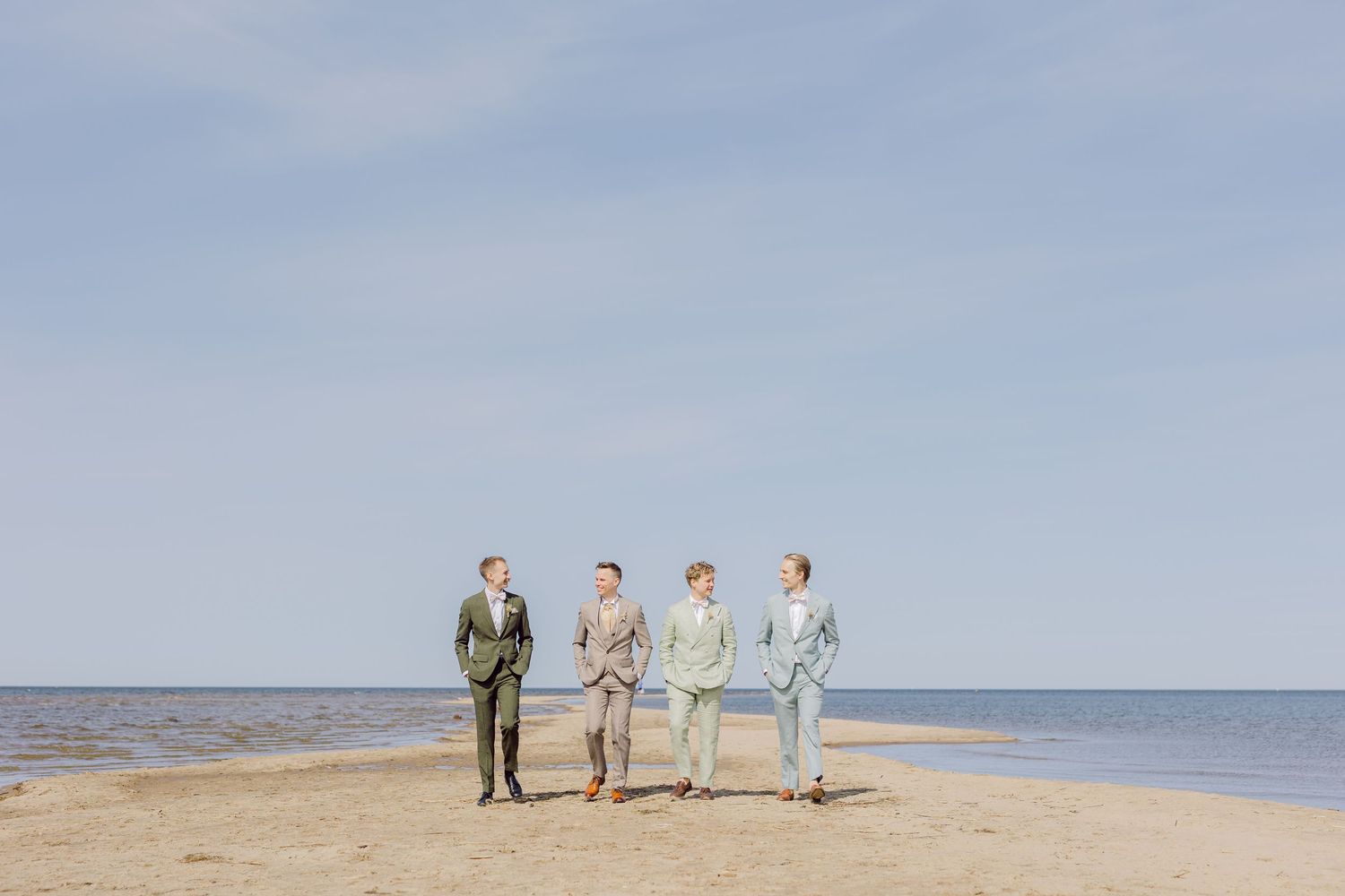 Four people walk together along a sandy beach shoreline under a soft blue sky on a calm day.