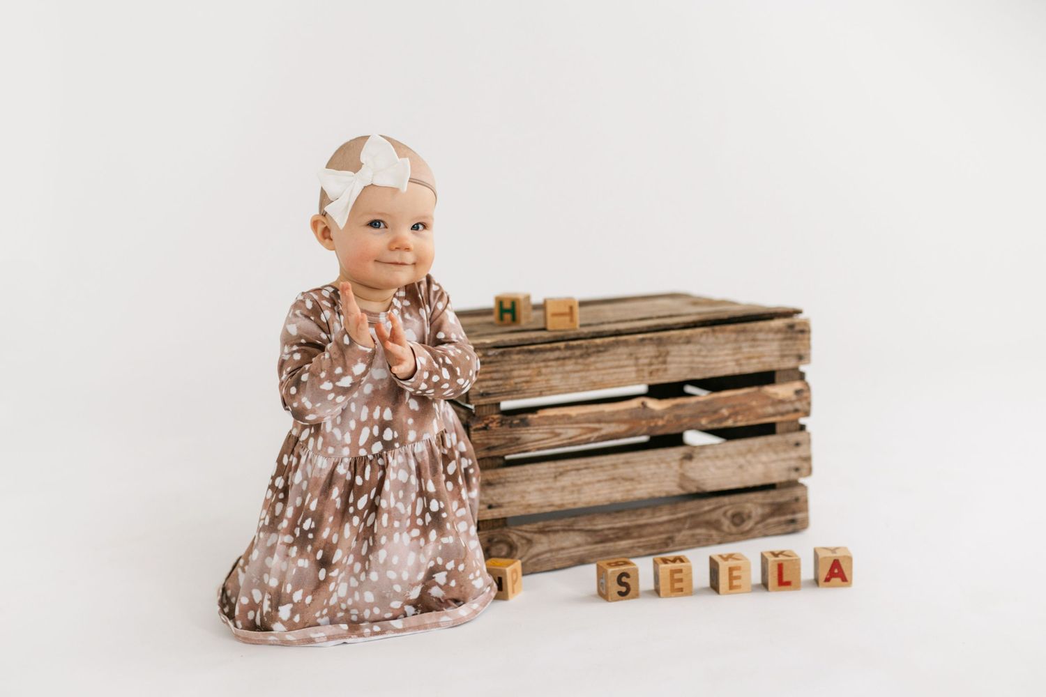 A baby in a floral dress poses next to vintage wooden crates and alphabet blocks.