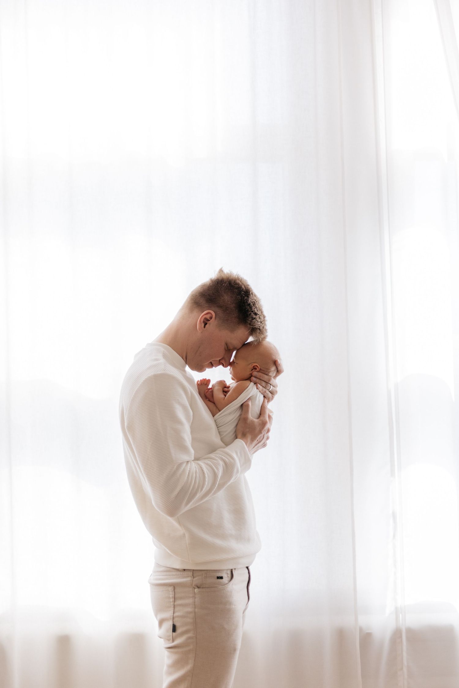 Parent in white sweater tenderly holding newborn baby against bright window background.