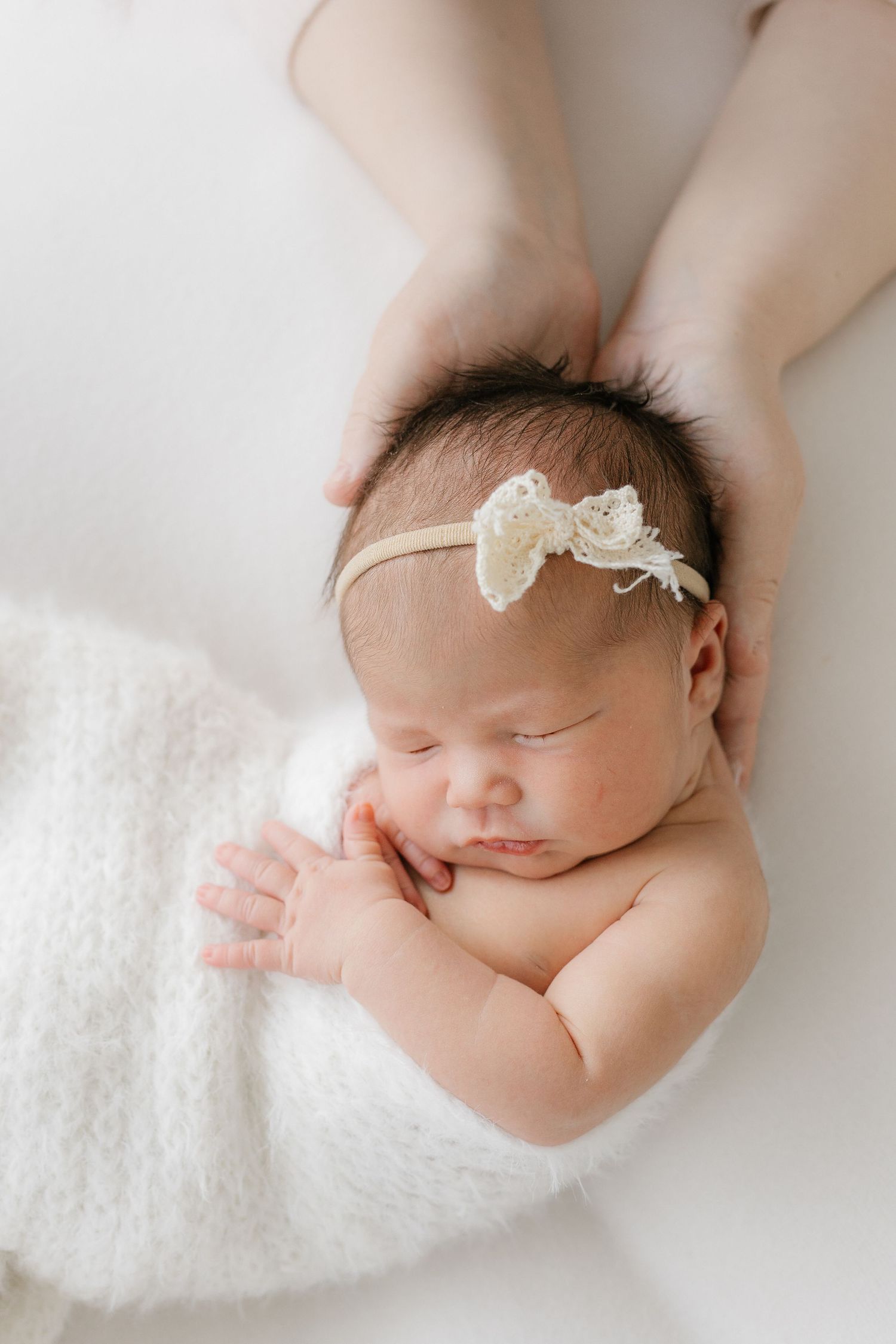 Sleeping newborn baby girl wearing a delicate lace bow headband, wrapped in a soft white blanket.