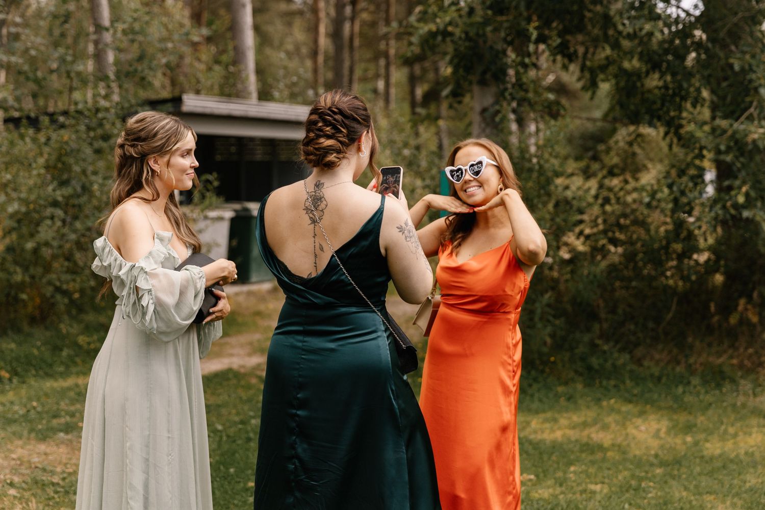 Three wedding guests in formal dresses enjoying drinks outdoors in natural setting.