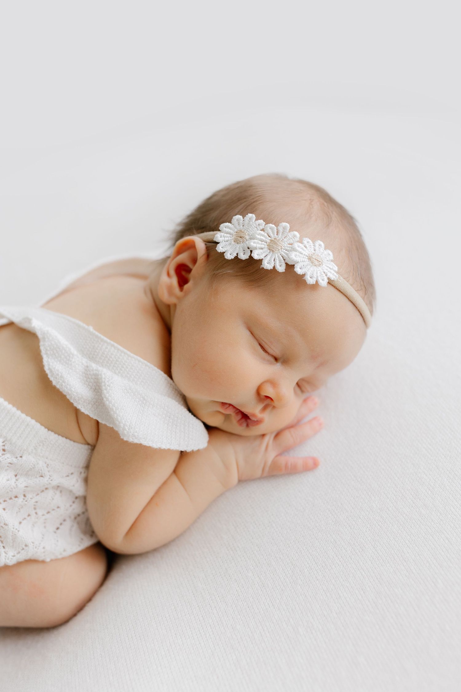 Sleeping newborn baby in white lace wrap and delicate floral headband lying on soft white backdrop.