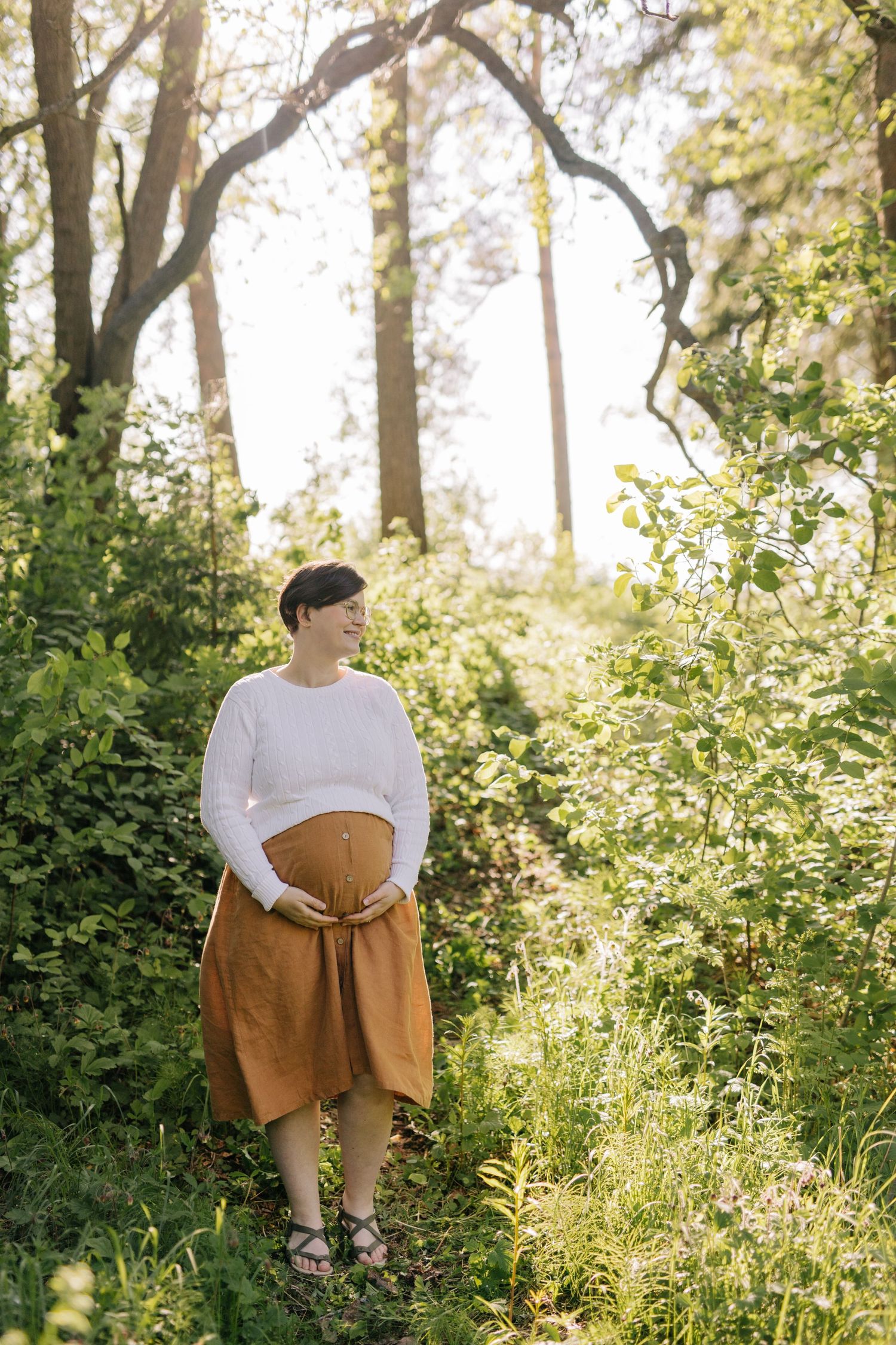 Maternity portrait in green forest setting showing pregnant belly in brown skirt.