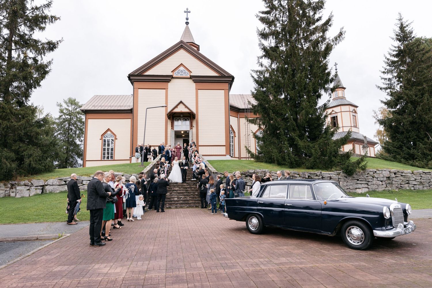Wedding party gathered outside traditional church with vintage black car on brick path.