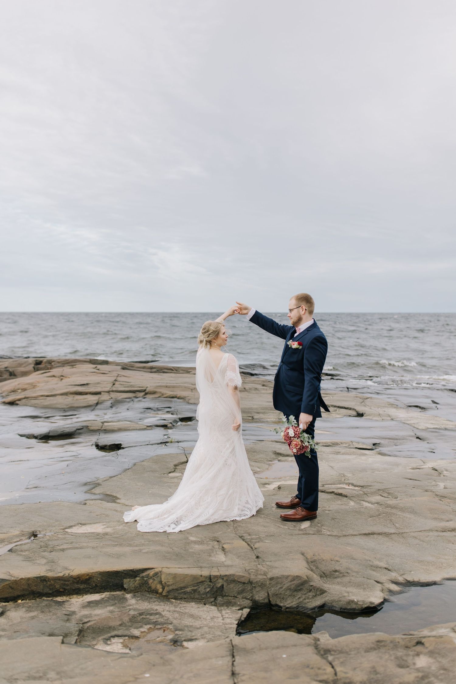 A romantic seaside wedding moment captured on rocky coastal cliffs under an overcast sky.