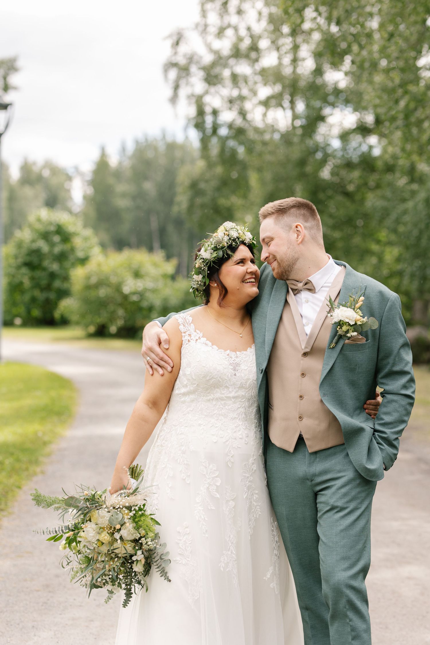 A couple in wedding attire share a joyful moment outdoors, with the bride wearing a floral crown and holding a green bouquet.
