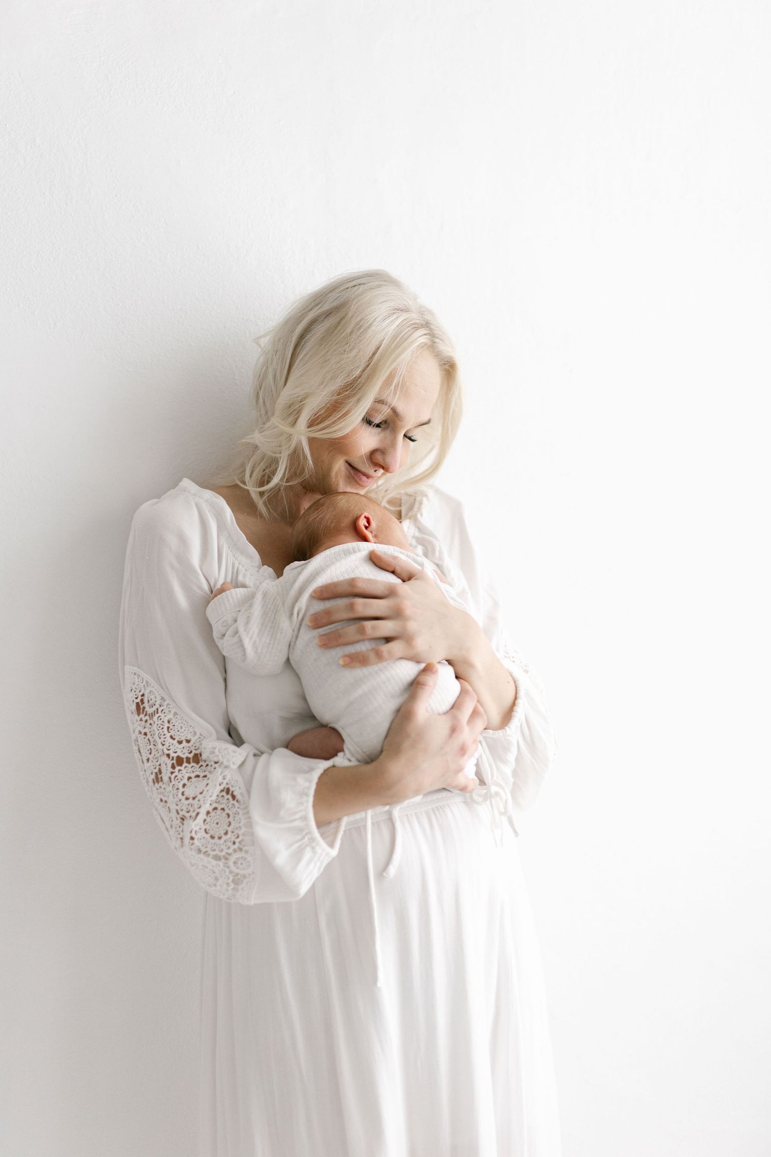 Mother in white lace dress holding newborn against white wall.