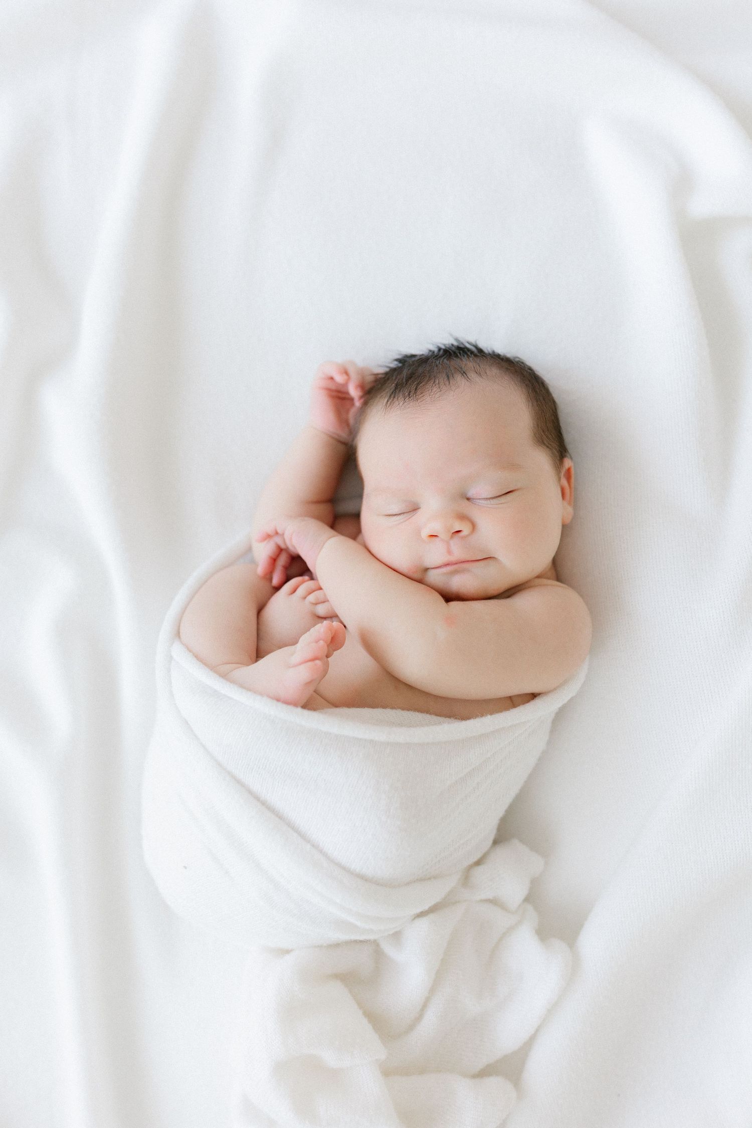 A peaceful sleeping newborn wrapped in soft white fabric against white bedding.