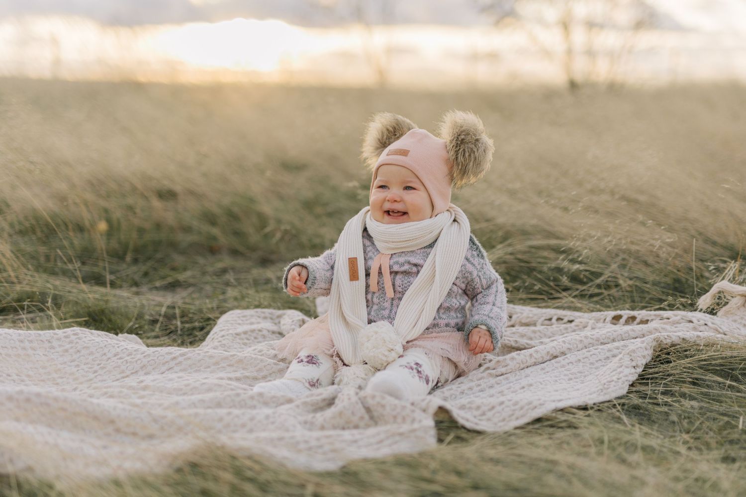 A laughing baby wearing winter clothes sits on blanket in grassy field.