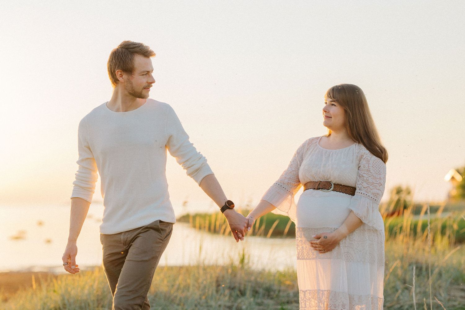 Expecting couple walking hand in hand during golden hour by the shore.