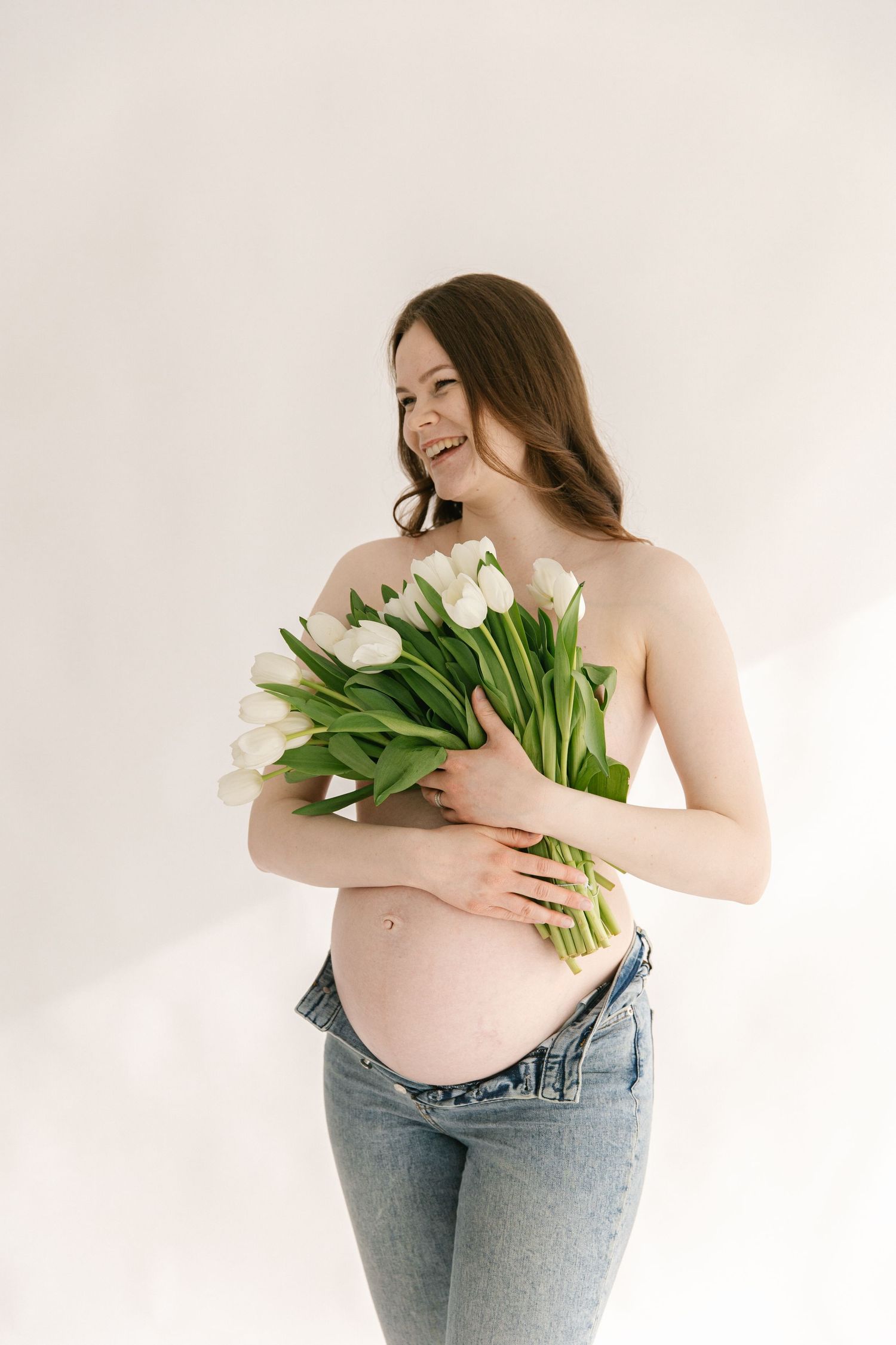 Pregnant woman in jeans holds white tulips against white background in artistic maternity photo series.