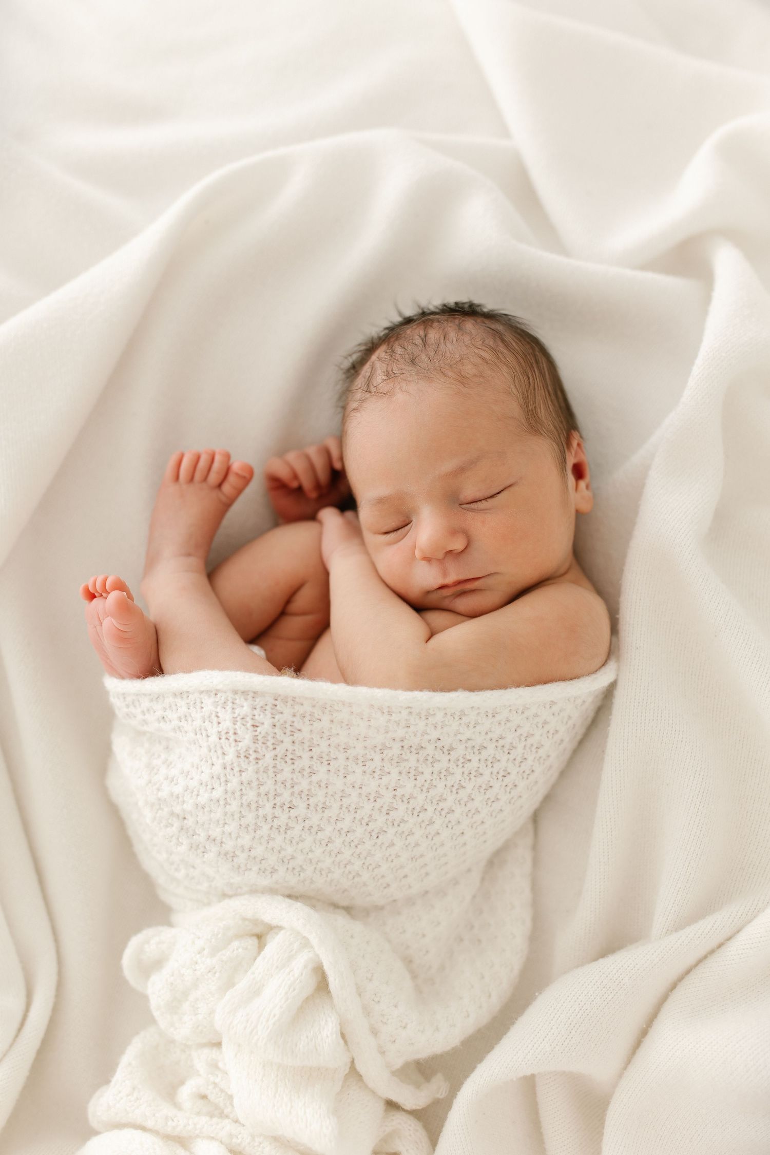 Peaceful newborn baby sleeping wrapped in a soft white knit blanket, curled in a natural fetal position.