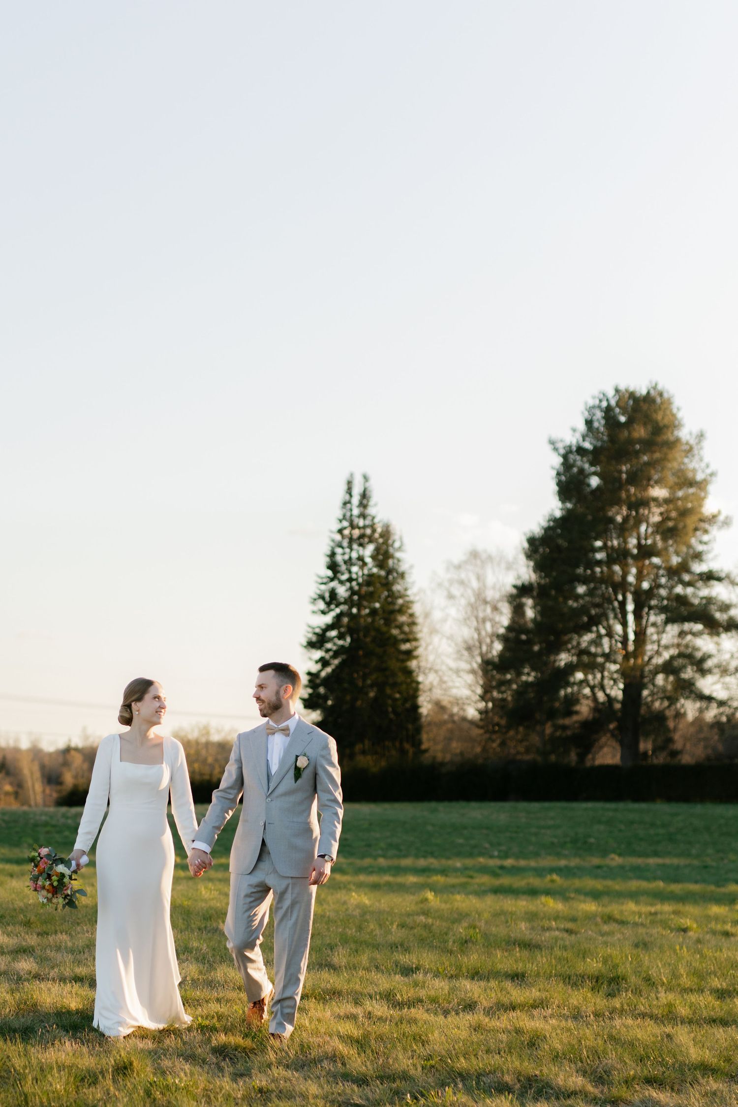 Newlywed couple walking hand in hand across a sunlit meadow with evergreen trees in the background.
