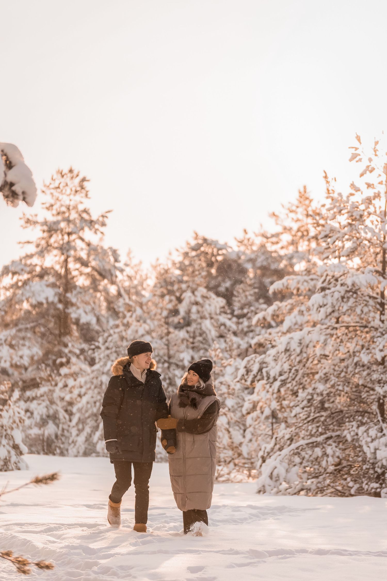 Couple walking together through snowy winter forest with warm sunset glow.