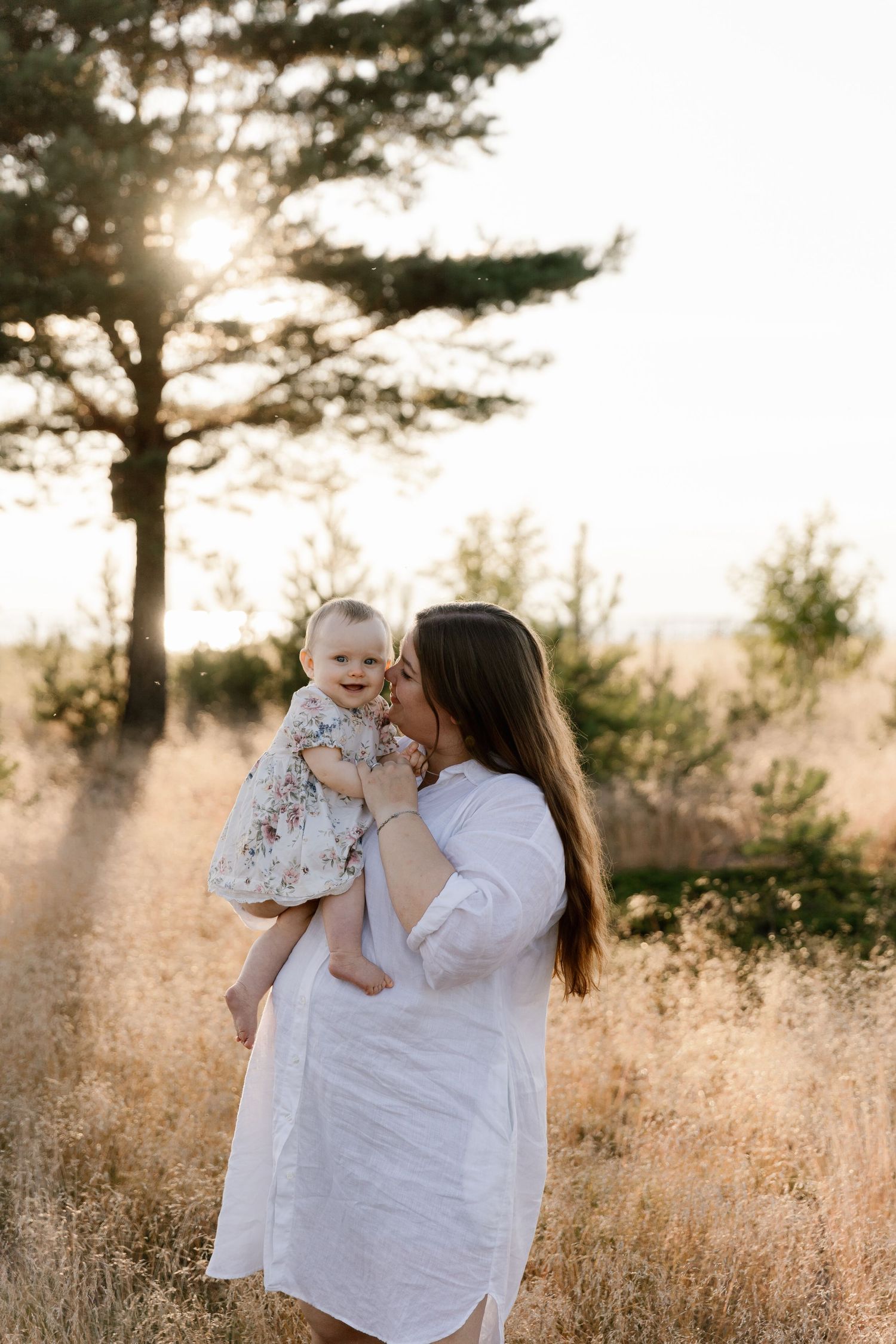 Mother in white dress holding baby outdoors with sunlight filtering through pine trees at sunset in golden field.
