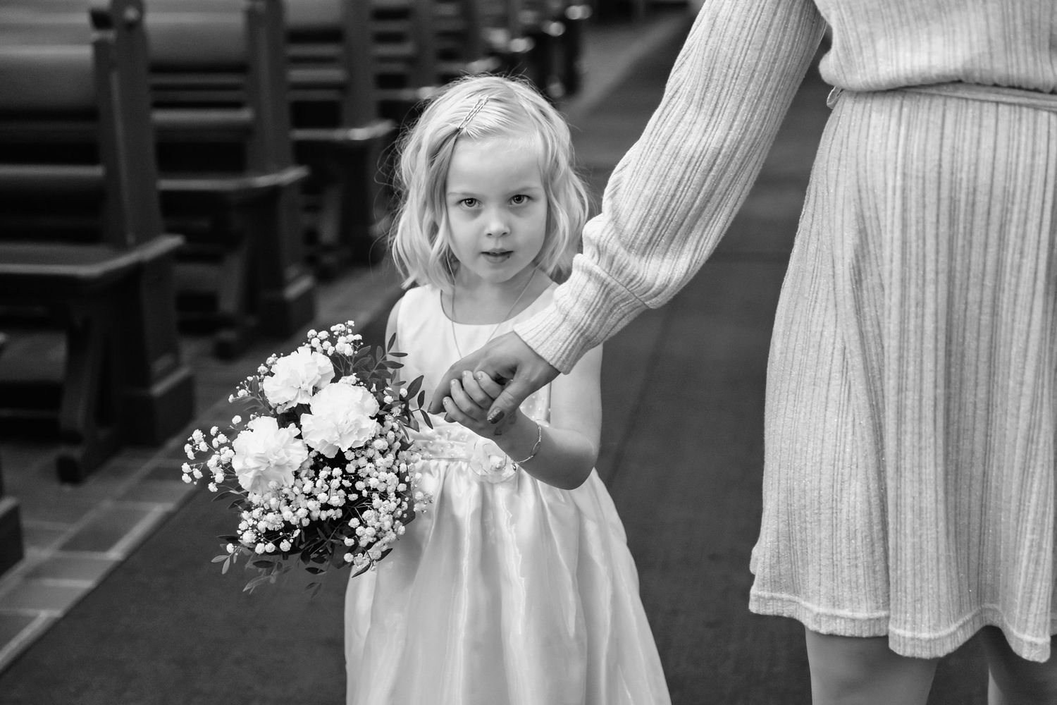 Young flower girl in white dress holding bouquet while walking down church aisle.