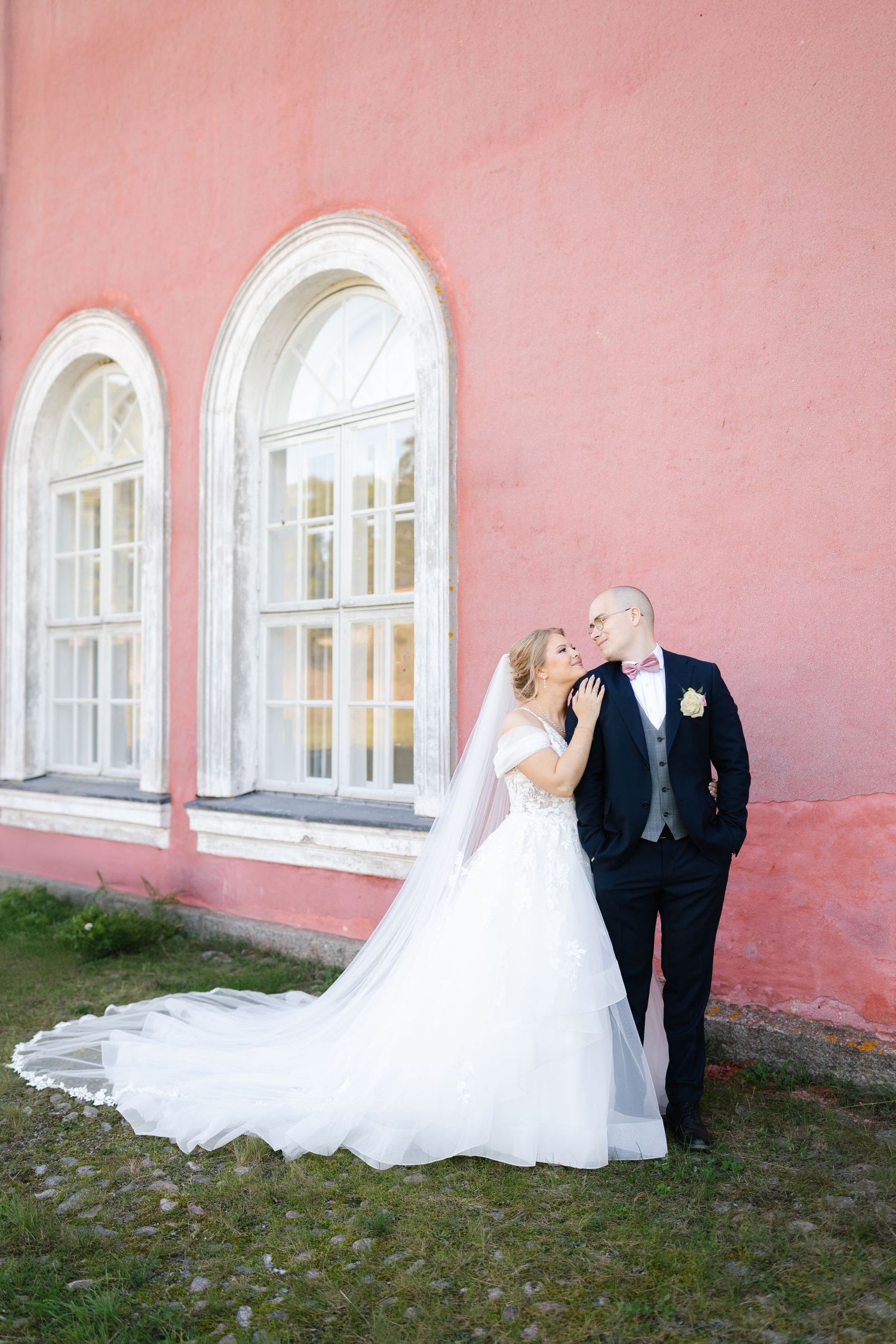 Wedding couple poses against pink wall with arched windows in elegant formal attire.