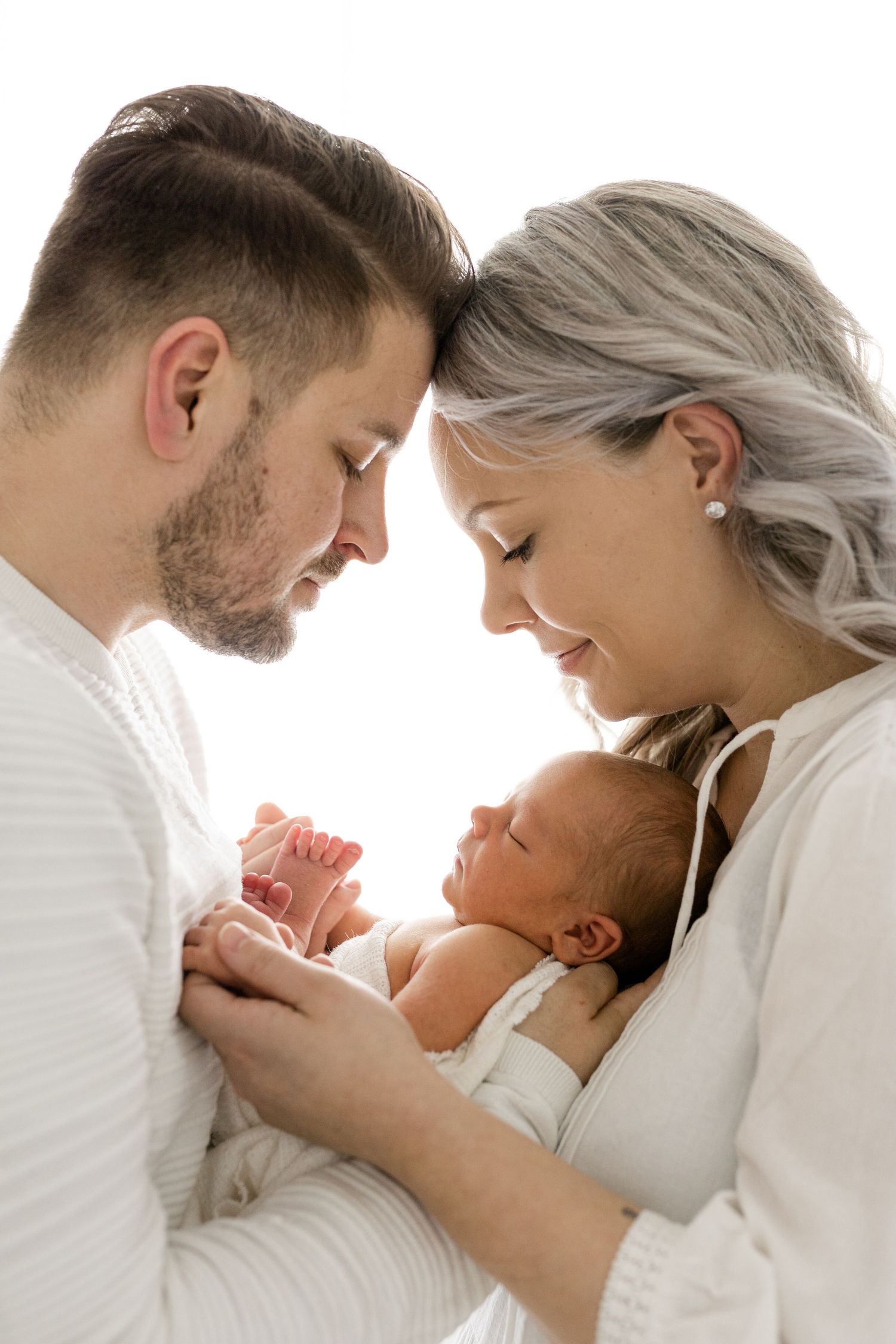 A peaceful newborn photoshoot with parents in white clothing against bright background.