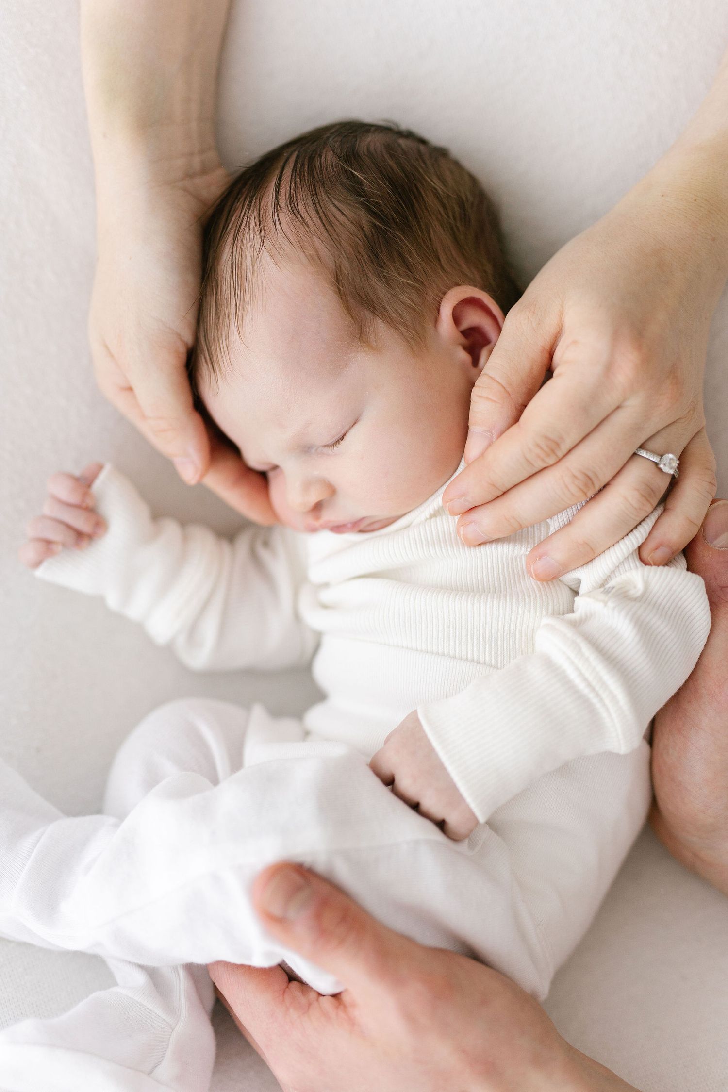 Gentle hands holding sleeping infant in white onesie.