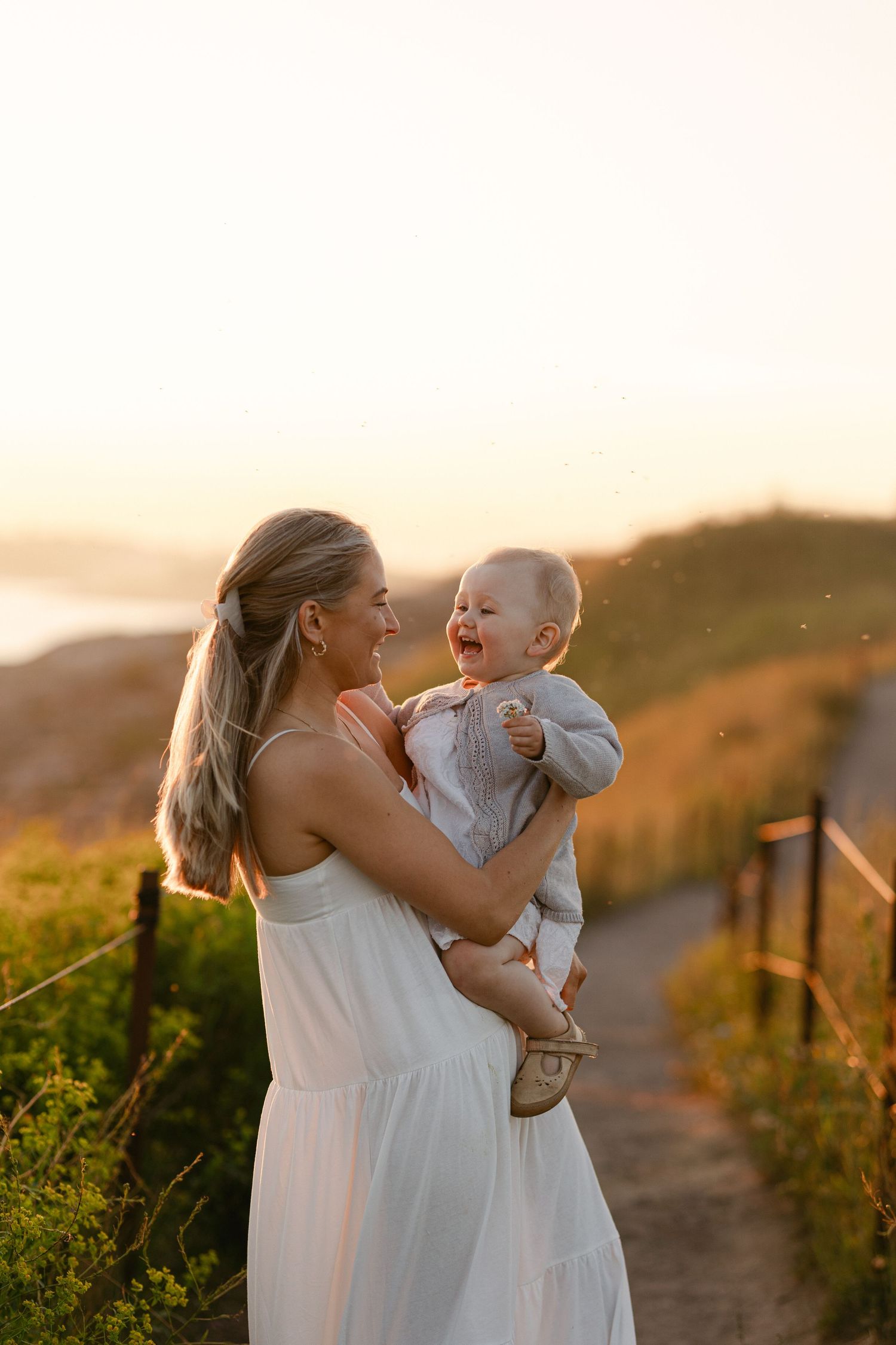 A heartwarming sunset moment captured on a hilltop path with a flowing white dress and joyful laughter.