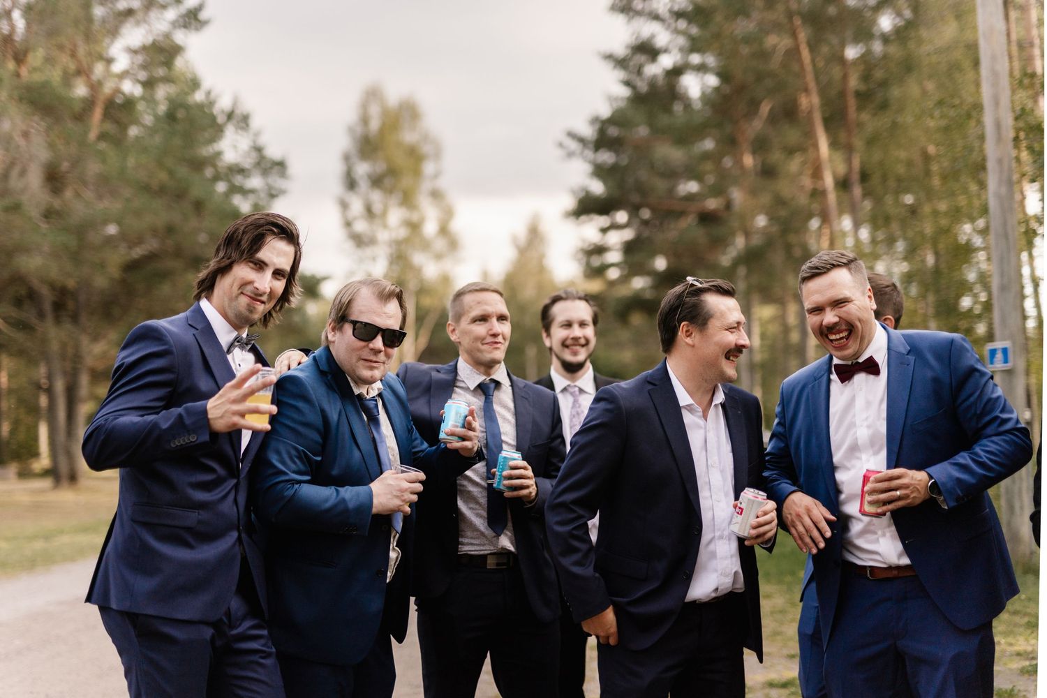 Groomsmen in formal suits sharing drinks and laughs outdoors in a pine forest setting.