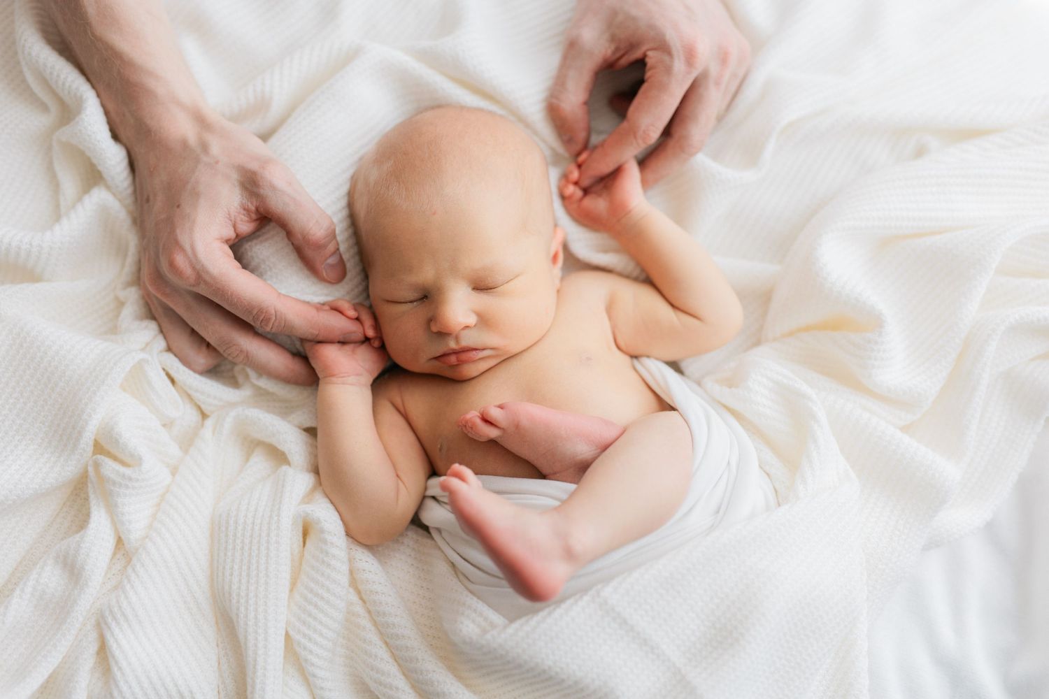 A peaceful newborn resting on white bedding while gentle hands touch their head.
