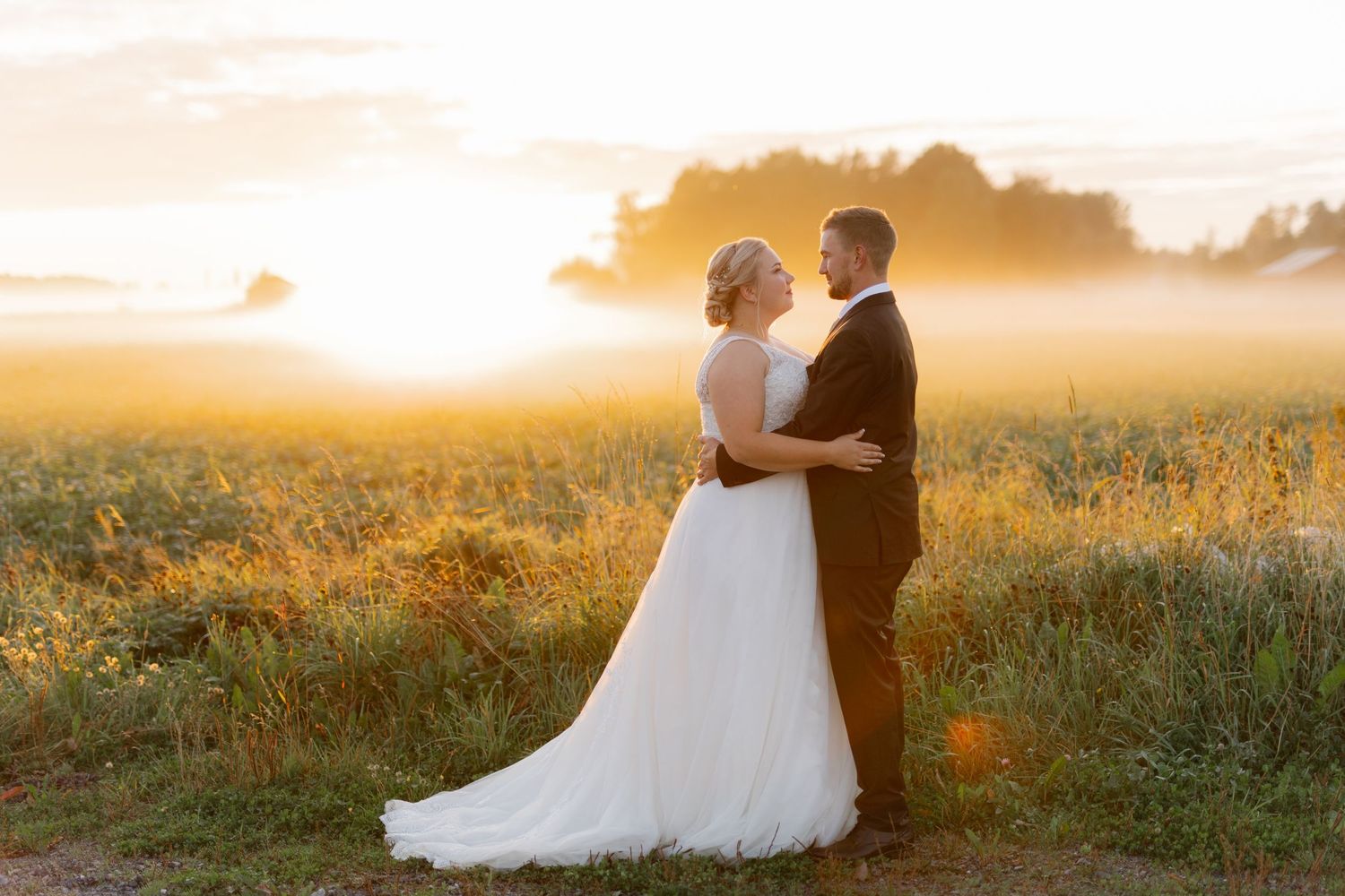 A couple in wedding attire embraces during golden hour sunset in a dreamy meadow field.