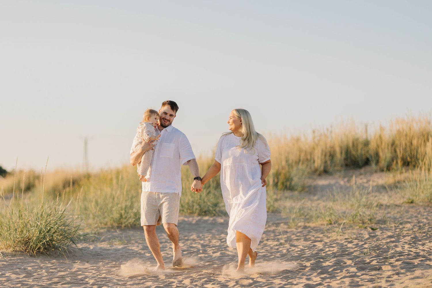 Family walking together on sandy beach at sunset wearing casual white outfits.