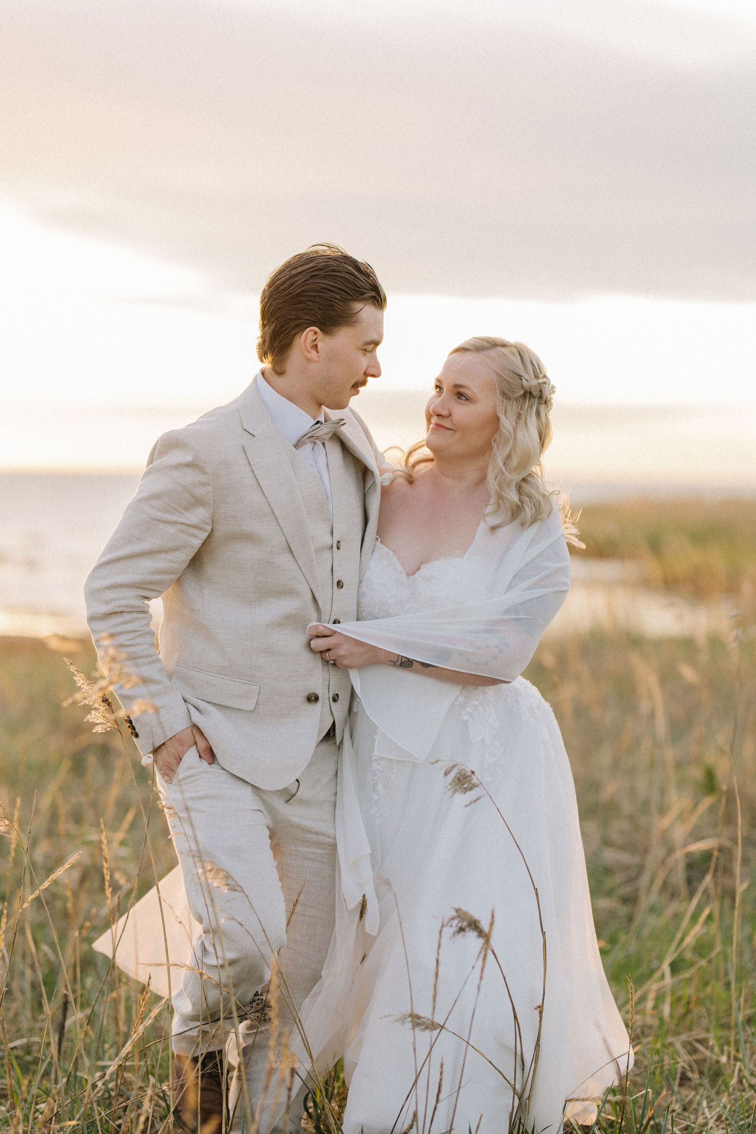 Wedding couple in light colored attire share tender moment in golden field at sunset.