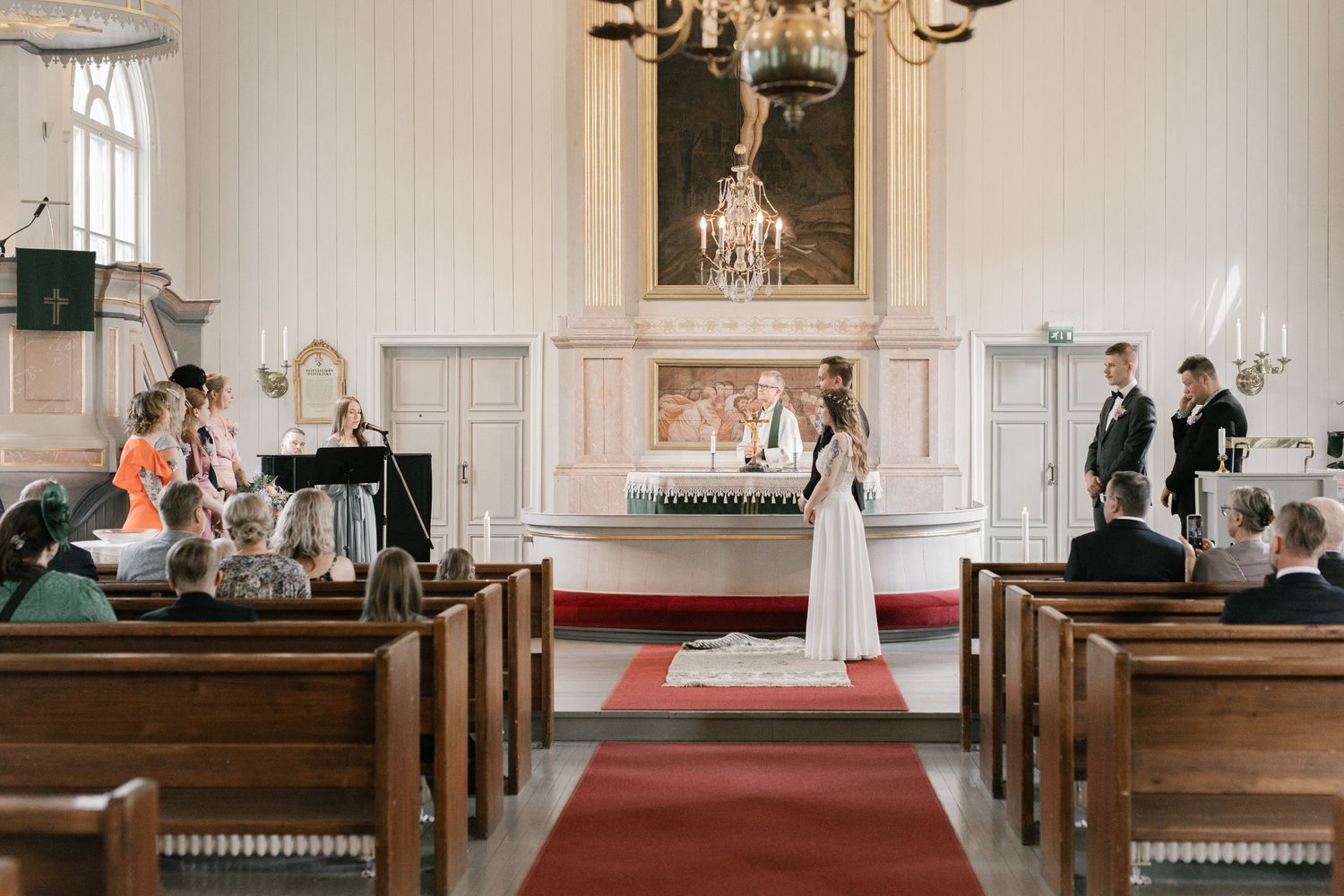 A wedding ceremony in progress inside a historic white chapel with traditional wooden pews and red carpet.