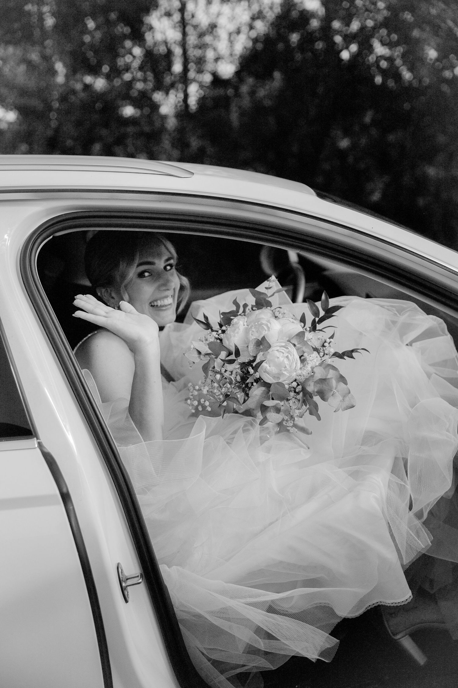 Black and white photo of bride with bouquet sitting in car wearing flowing white dress.
