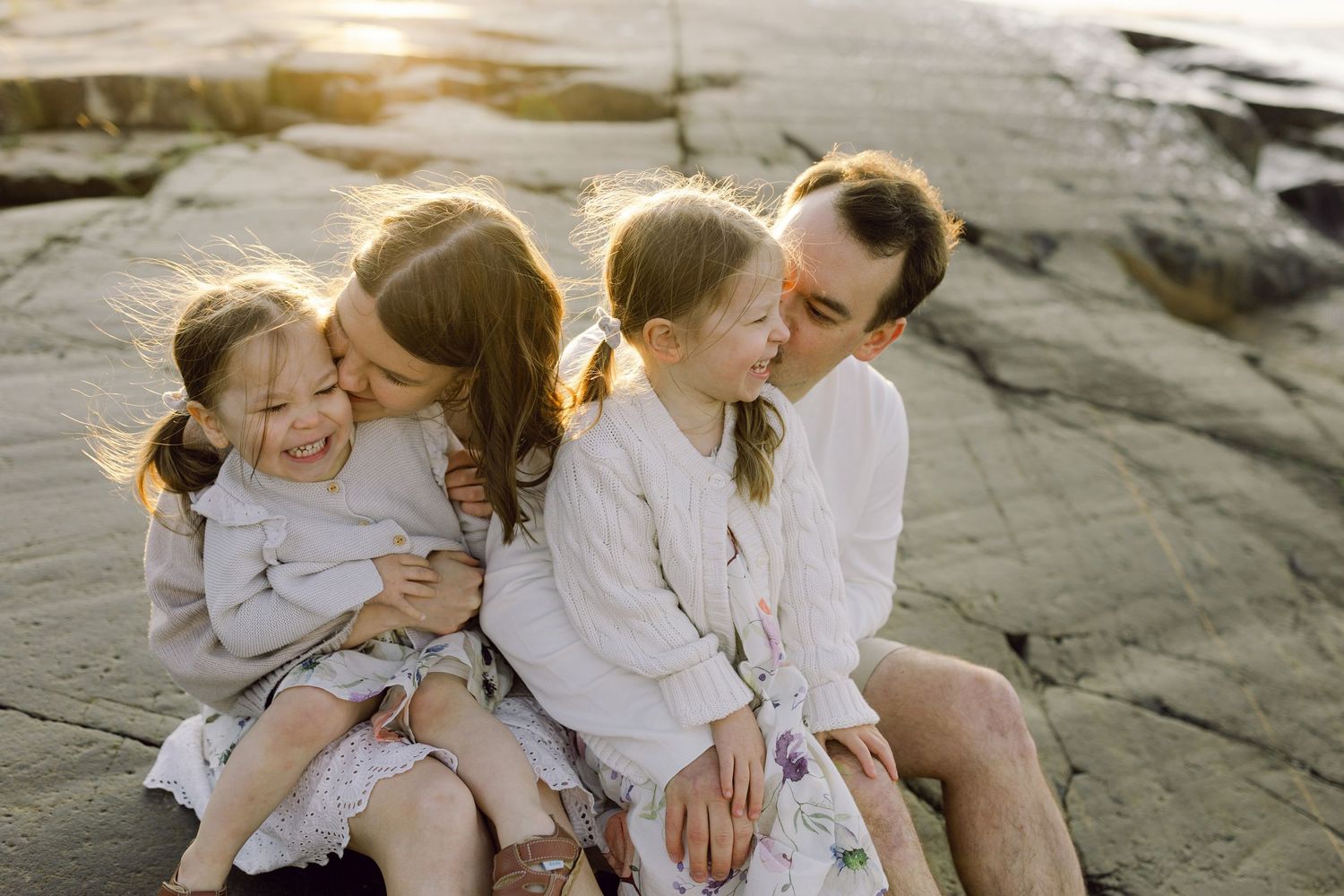 A family sits together on coastal rocks at golden hour wearing light colored clothing.