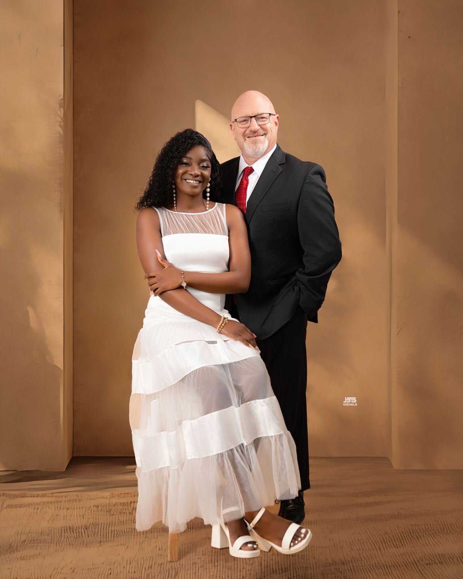 A couple poses together, woman in white tiered dress and man in black suit with red tie, against a tan background.