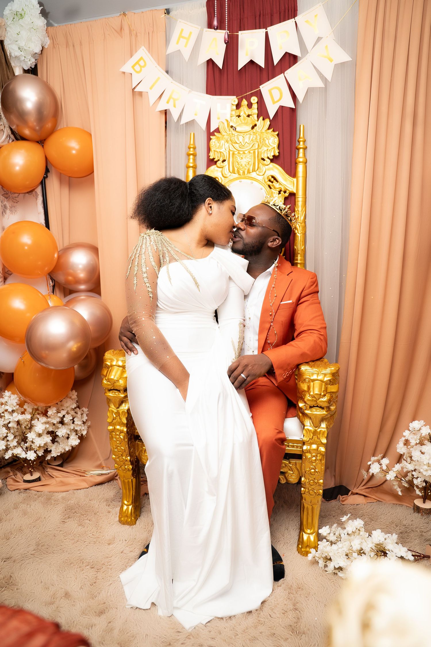 Couple sharing a kiss on gold throne at decorated birthday party with balloons.
