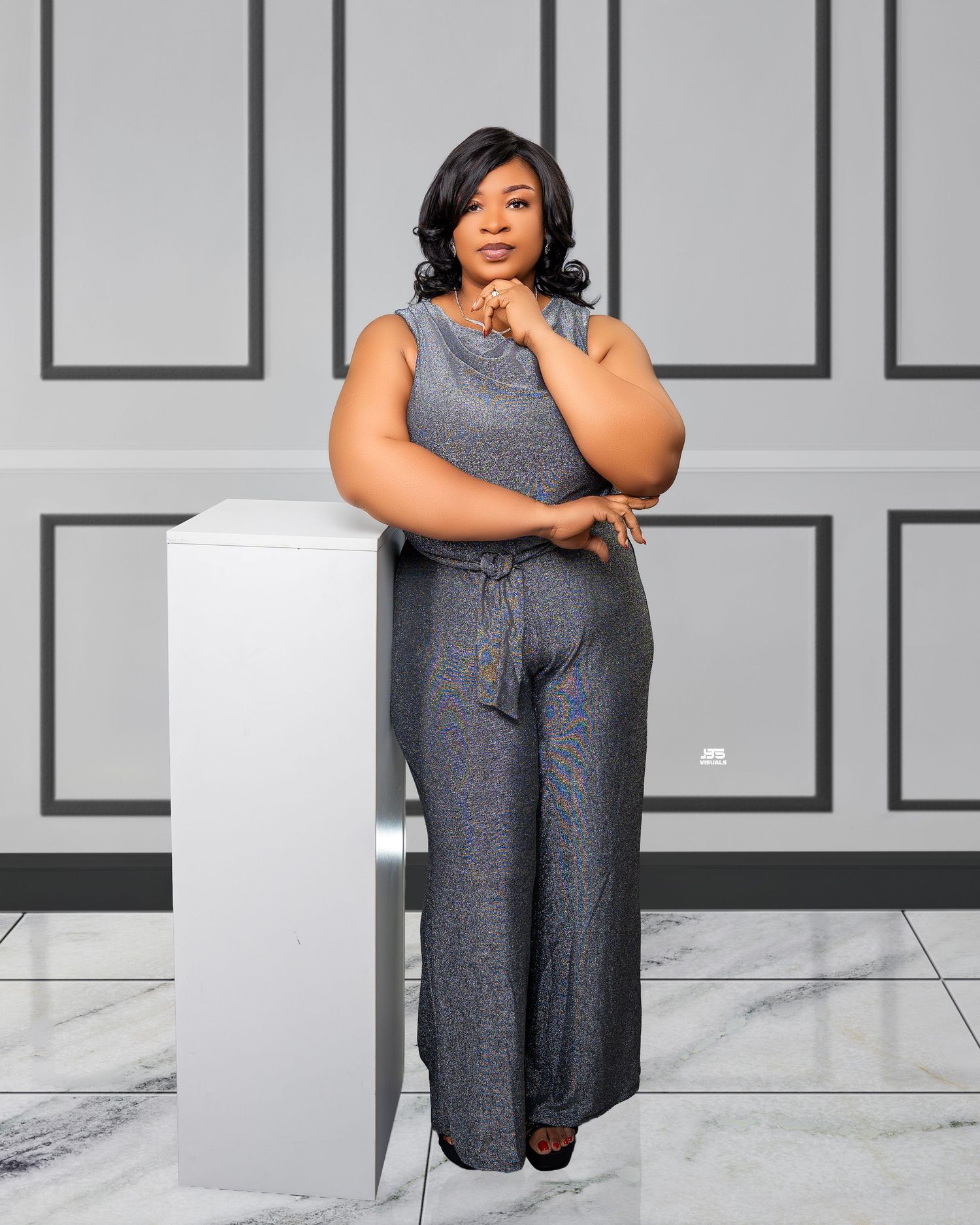 Confident plus-size woman poses in a sleeveless gray jumpsuit beside a white pedestal in a studio.