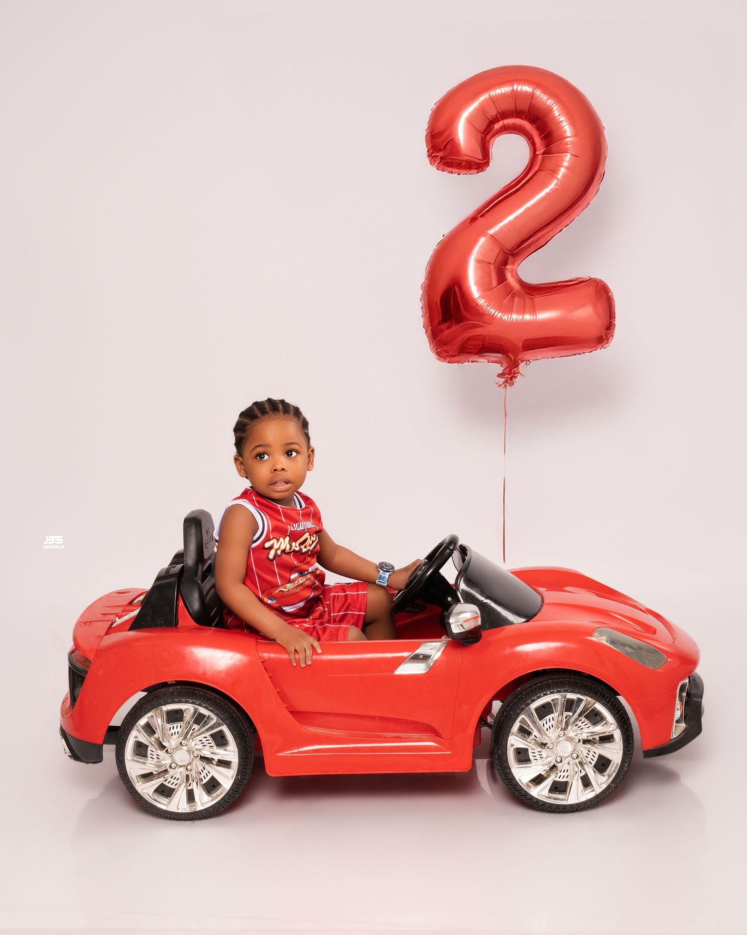 Young boy in red outfit sits in a red toy car beside a red number 2 balloon for birthday photos.