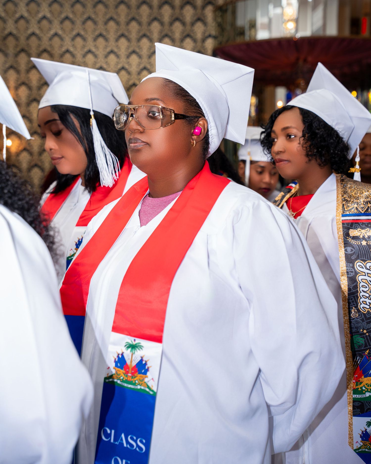 Graduate wearing white cap and gown with Haitian flag stole at graduation ceremony.