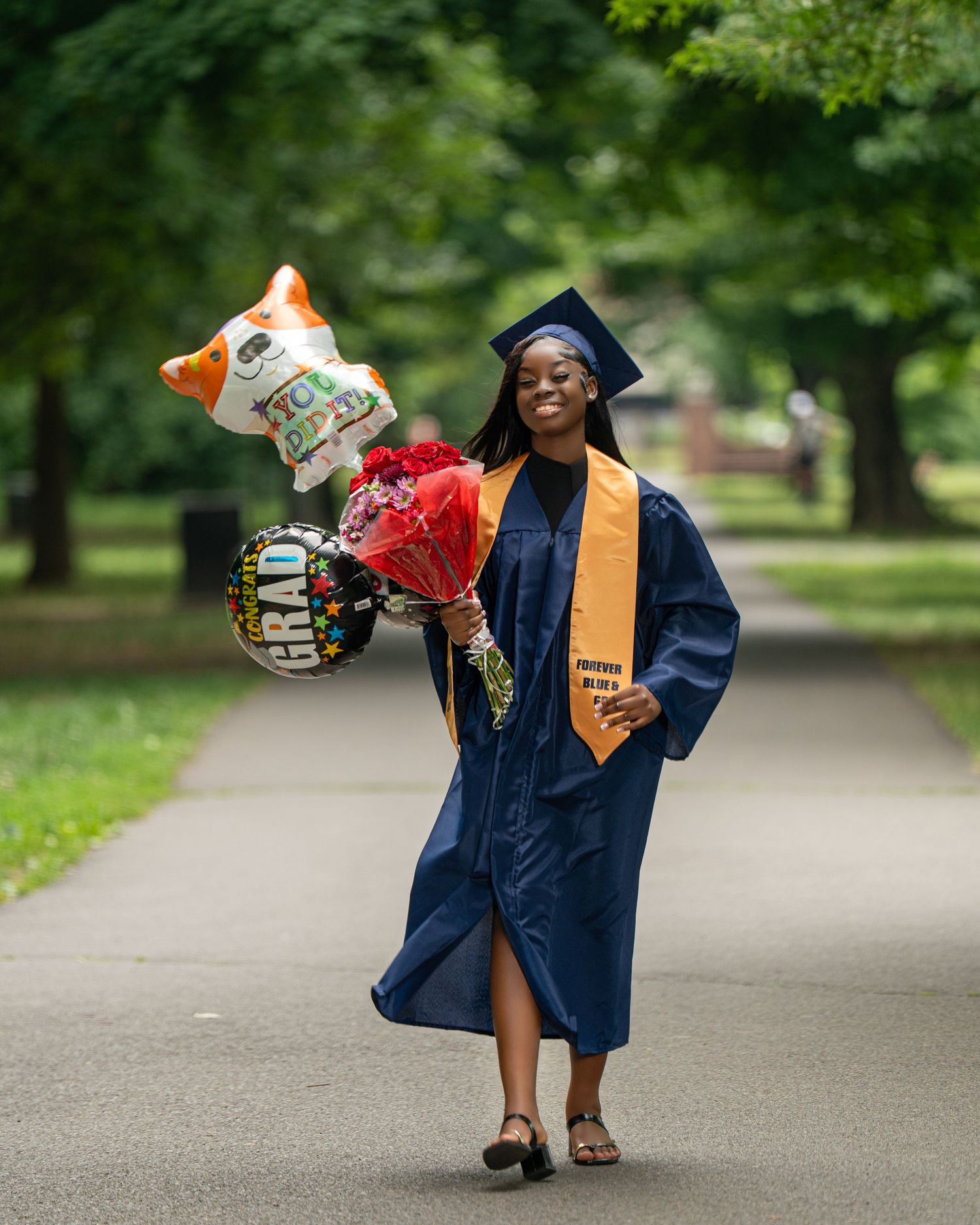 Graduate in navy blue cap and gown posing with colorful balloons outdoors.