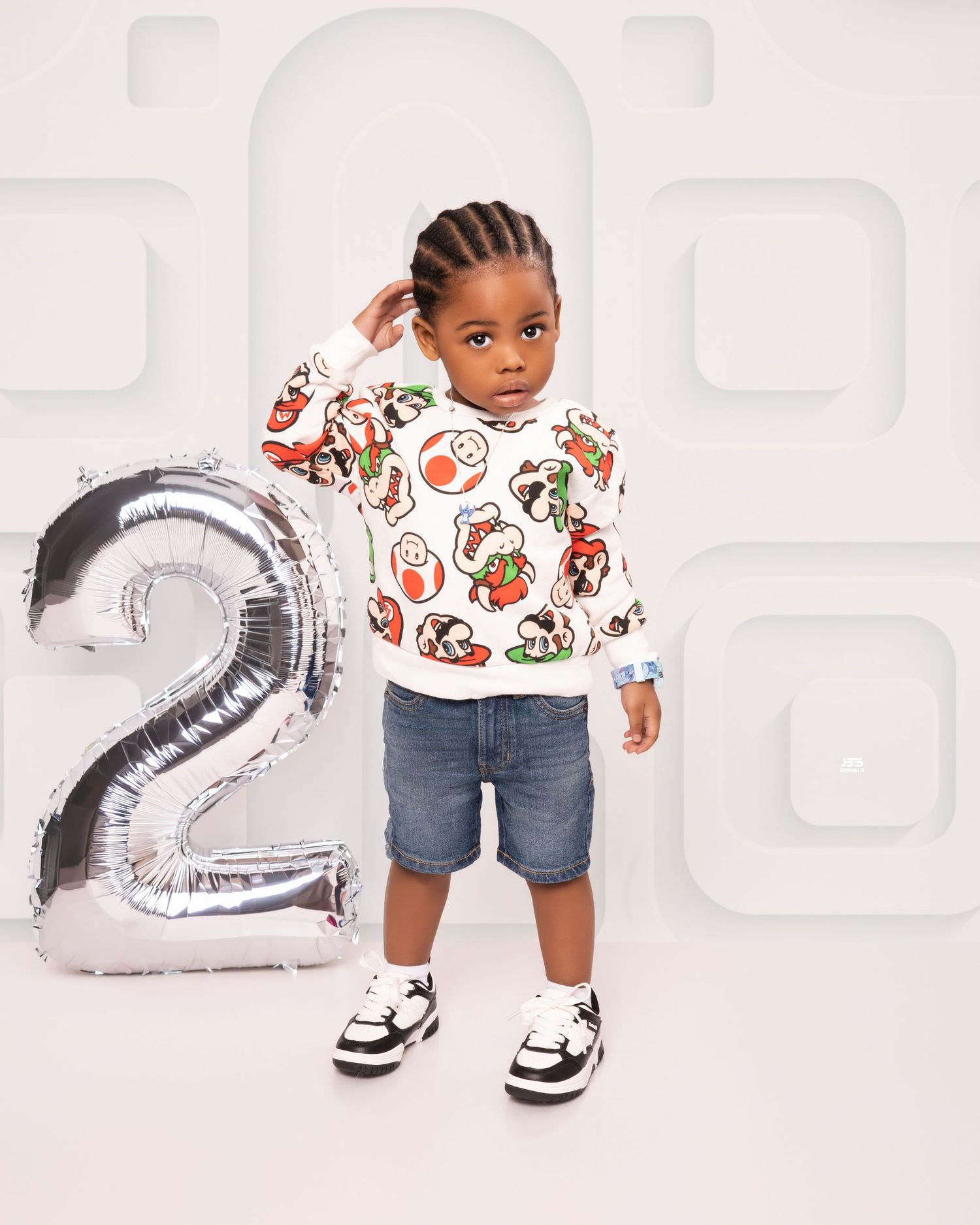 Stylish toddler boy in colorful graphic sweatshirt and denim shorts poses with silver number 2 balloon.